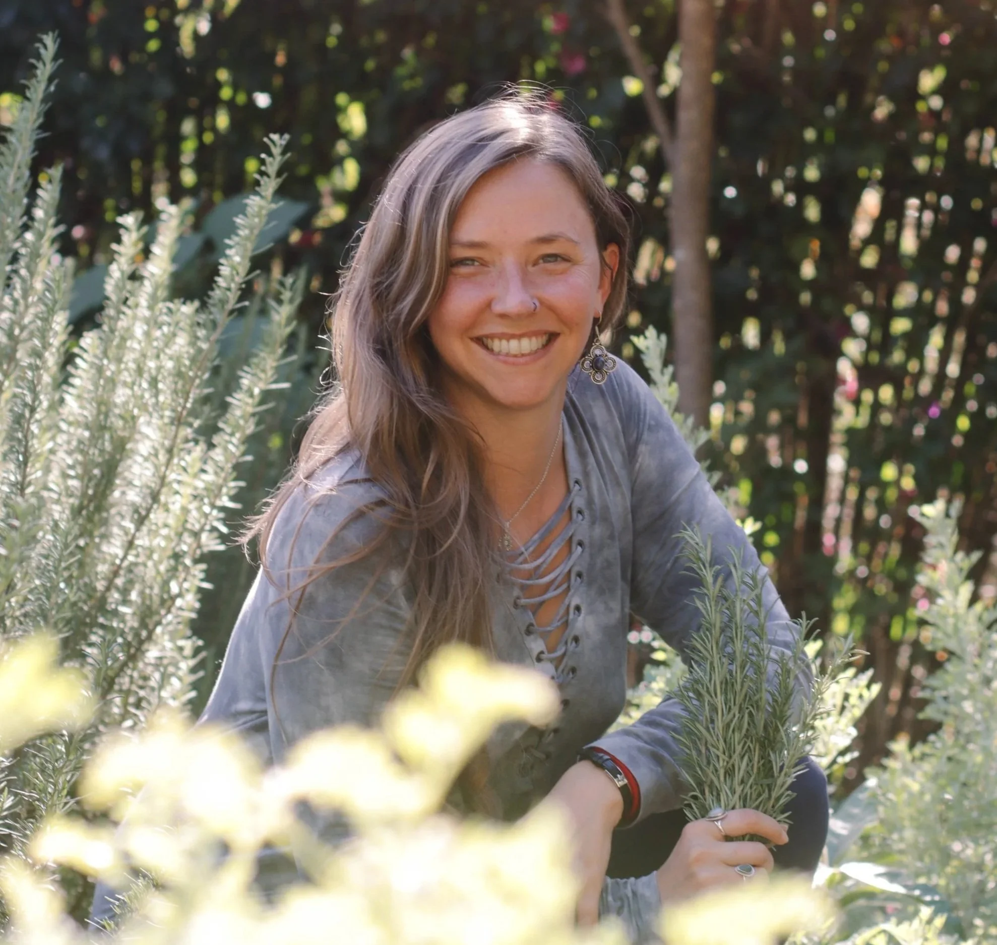 A smiling woman with long brown hair and earrings outdoors among greenery and plants, with sunlight filtering through trees in the background.