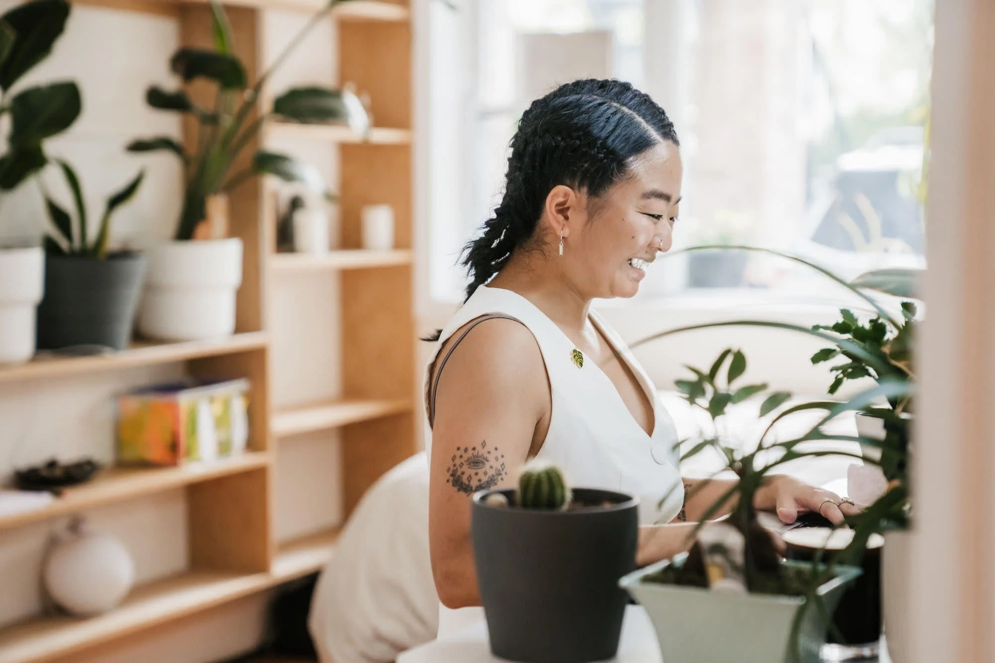 Woman sitting and smiling at a laptop, surrounded by indoor plants and shelves in a well-lit room.