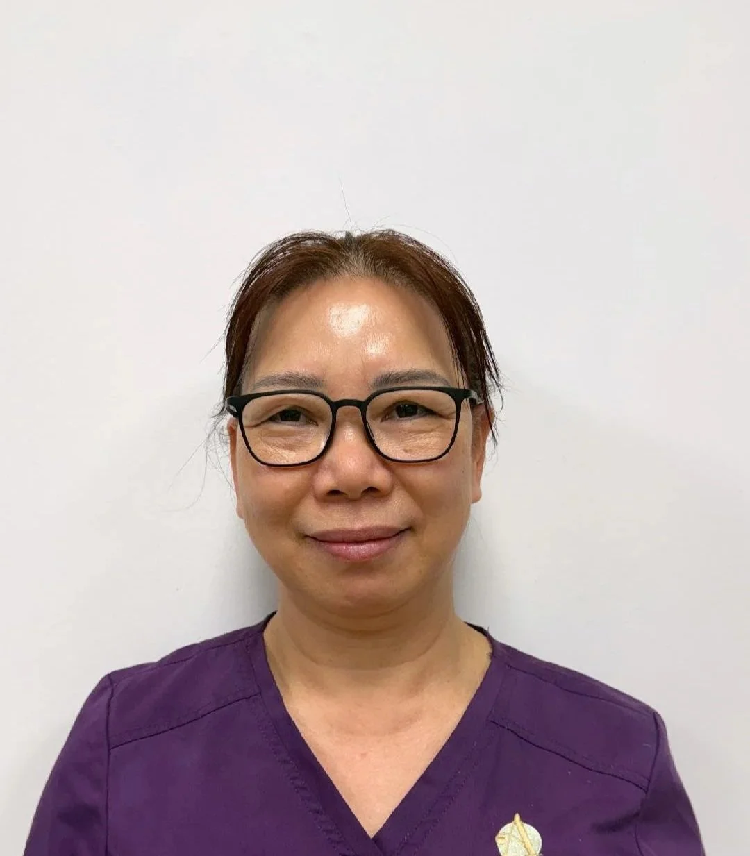 A massage therapist with short brown hair and glasses wearing a purple medical uniform in front of a plain white wall