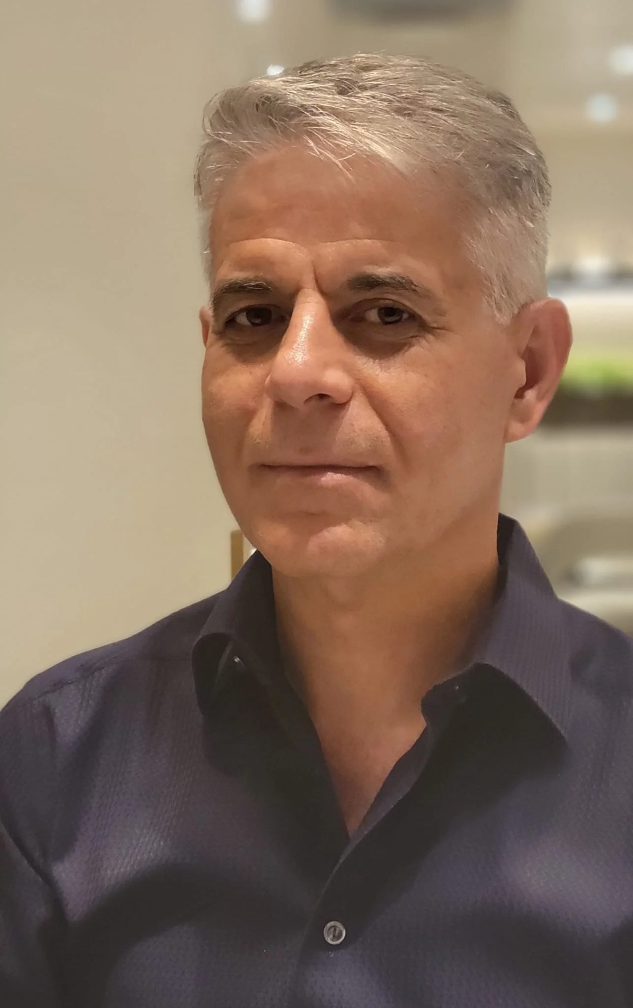 A middle-aged man with silver hair, wearing a dark button-up shirt, looking directly at the camera with a slight smile against a blurred indoor background.