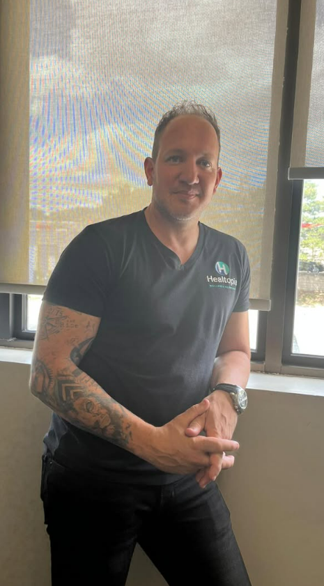 Man with short dark hair and a tattoo on his left arm, wearing a black T-shirt with a blue and white logo on the chest, standing against a plain beige wall.