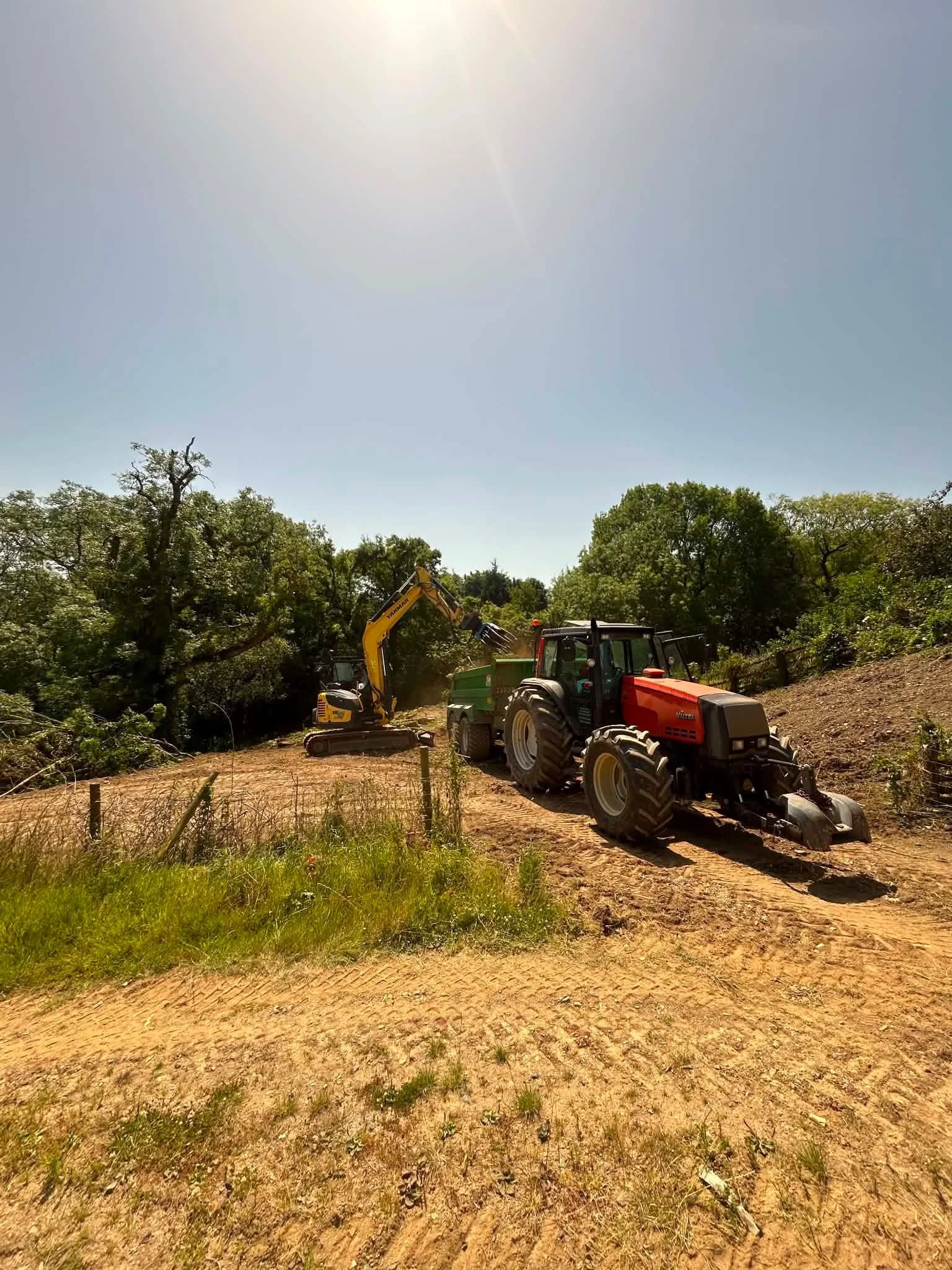 Construction site with a tractor and a small excavator working on the land, surrounded by trees under a bright sunny sky.