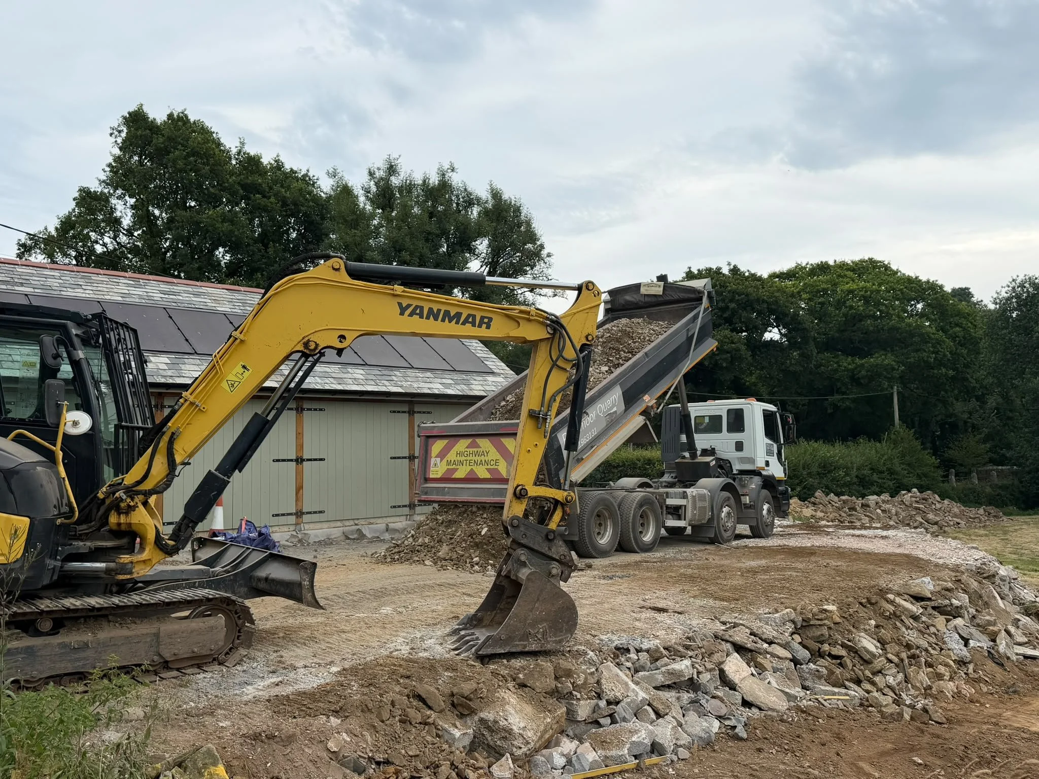 Construction site with a yellow Yanmar excavator moving dirt and rocks, and a dump truck with 'Highway Maintenance' written on its side, against cloudy sky and trees in the background.