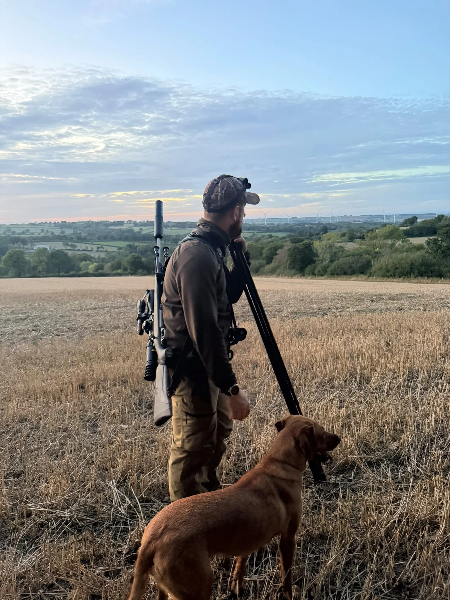 A man standing in a dry field with a brown dog, looking at the horizon as the sky changes color at sunset, with wind turbines in the distance.
