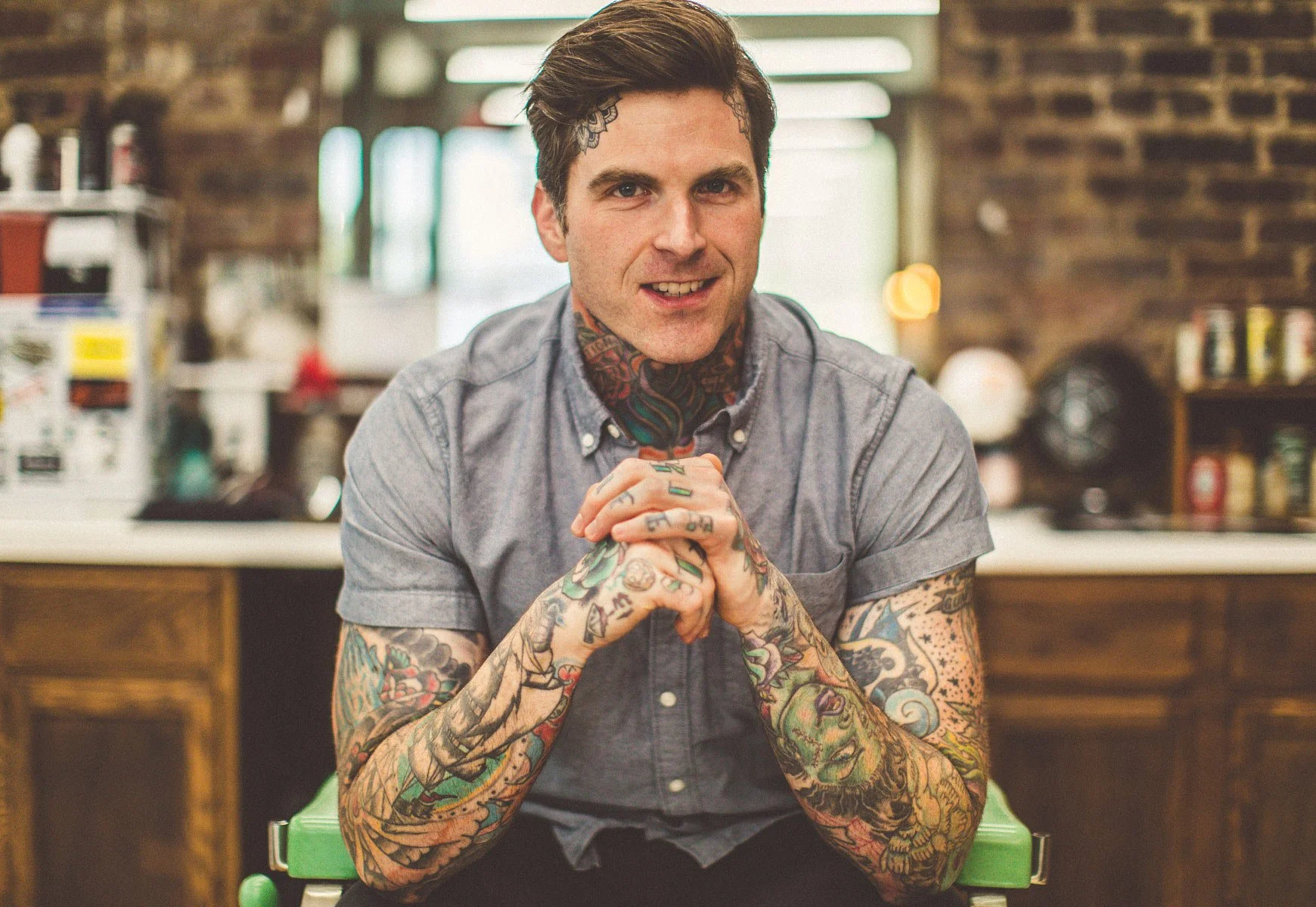A young man with dark hair, tattoos on his arms and neck, wearing a gray short-sleeved button-up shirt, sitting in a kitchen with brick walls and wooden cabinets, smiling and clasping his hands together.