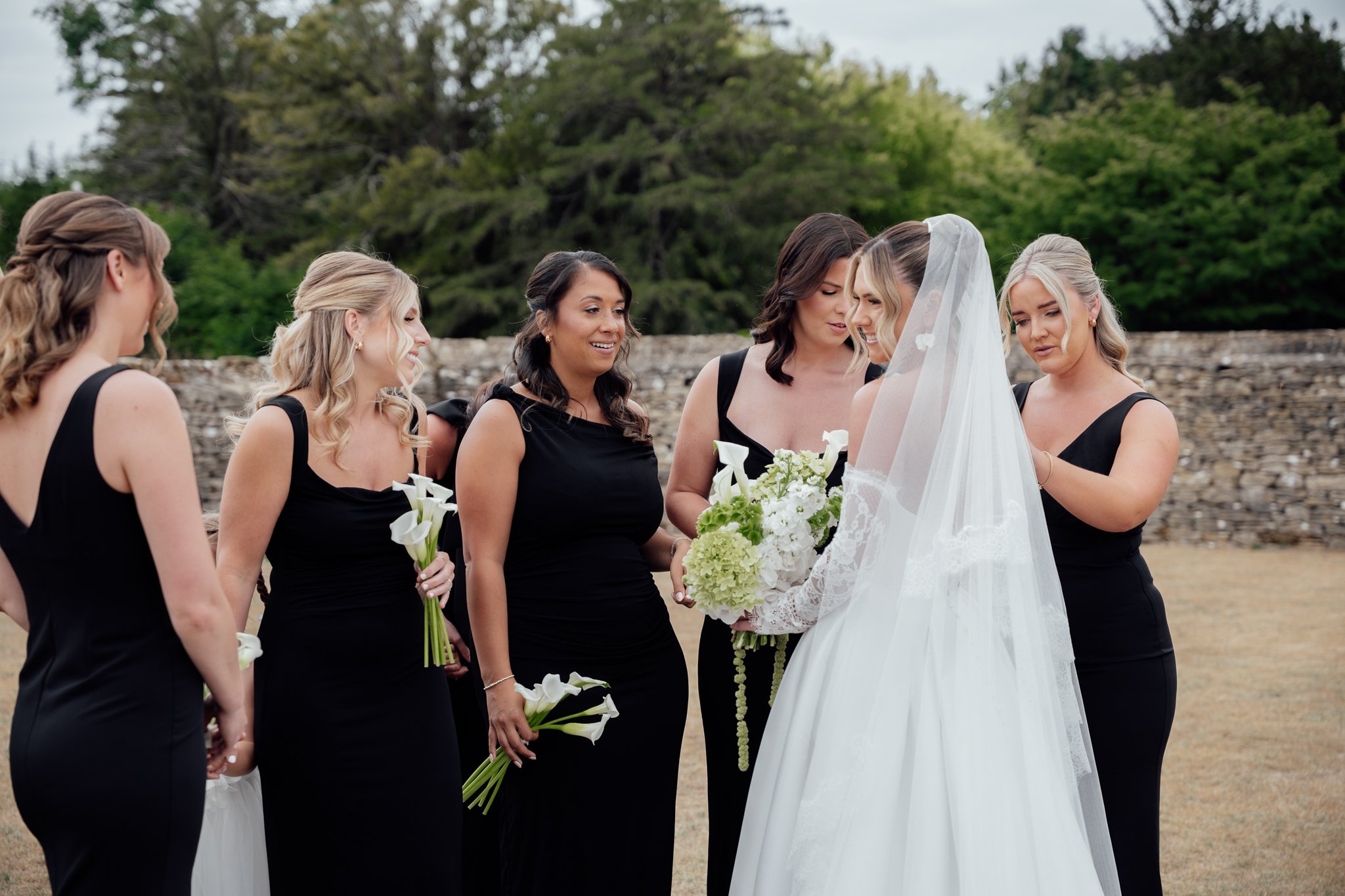 Bride in wedding dress with veil holding bouquet, surrounded by five bridesmaids in black dresses, outdoors with trees and stone wall in background.