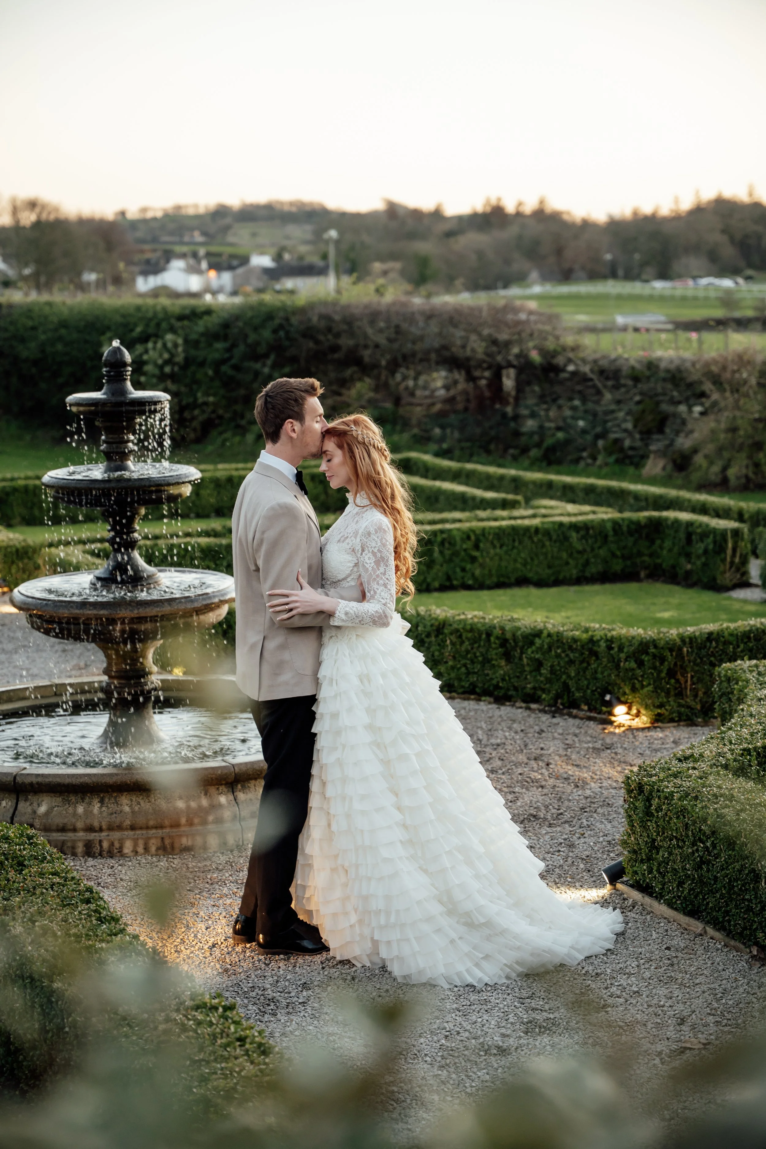 Bride and groom embracing by the fountain in the gardens at Cartmel Old Grammar, surrounded by manicured hedges and soft evening light.