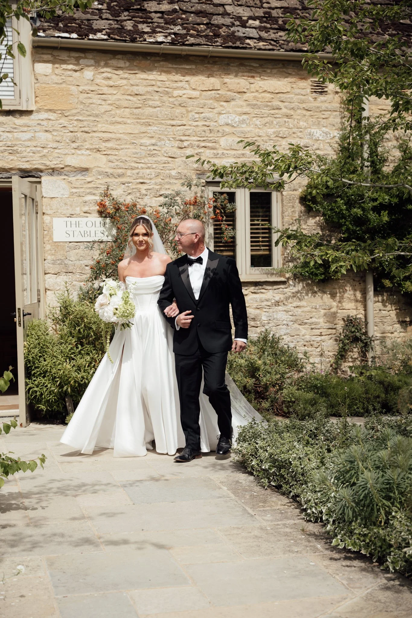 A bride walking arm in arm with her father at Caswell House, making her way toward the ceremony with the stone barn behind them.
