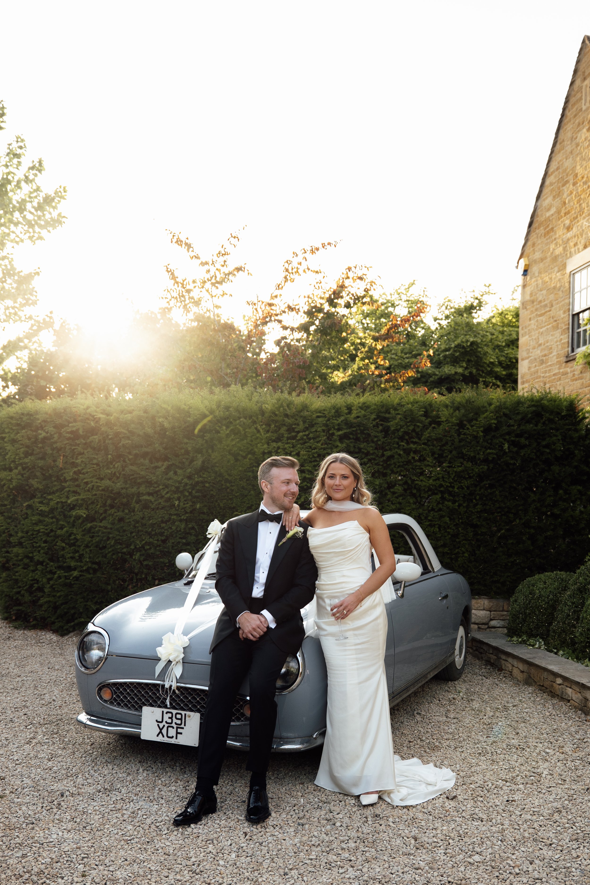 A newlywed couple in wedding attire standing in front of a vintage grey car decorated with white ribbons and flowers, outdoors during sunset at Charingworth House