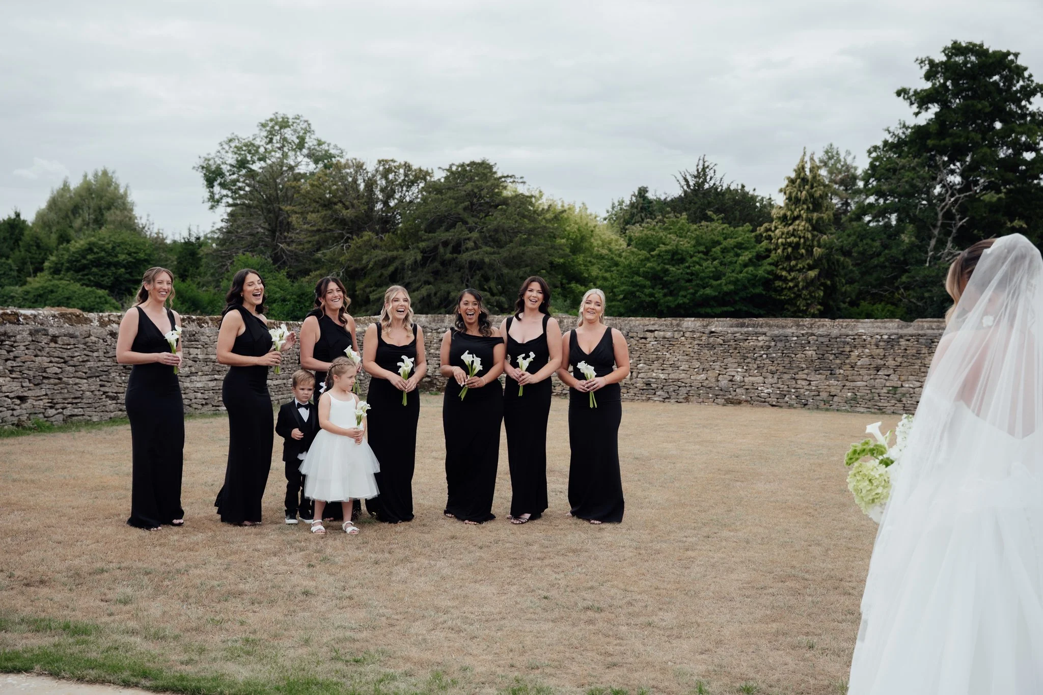 A bride in a white wedding dress and veil facing a group of seven women in black dresses and two children, holding flowers outdoors near a stone wall and green trees.