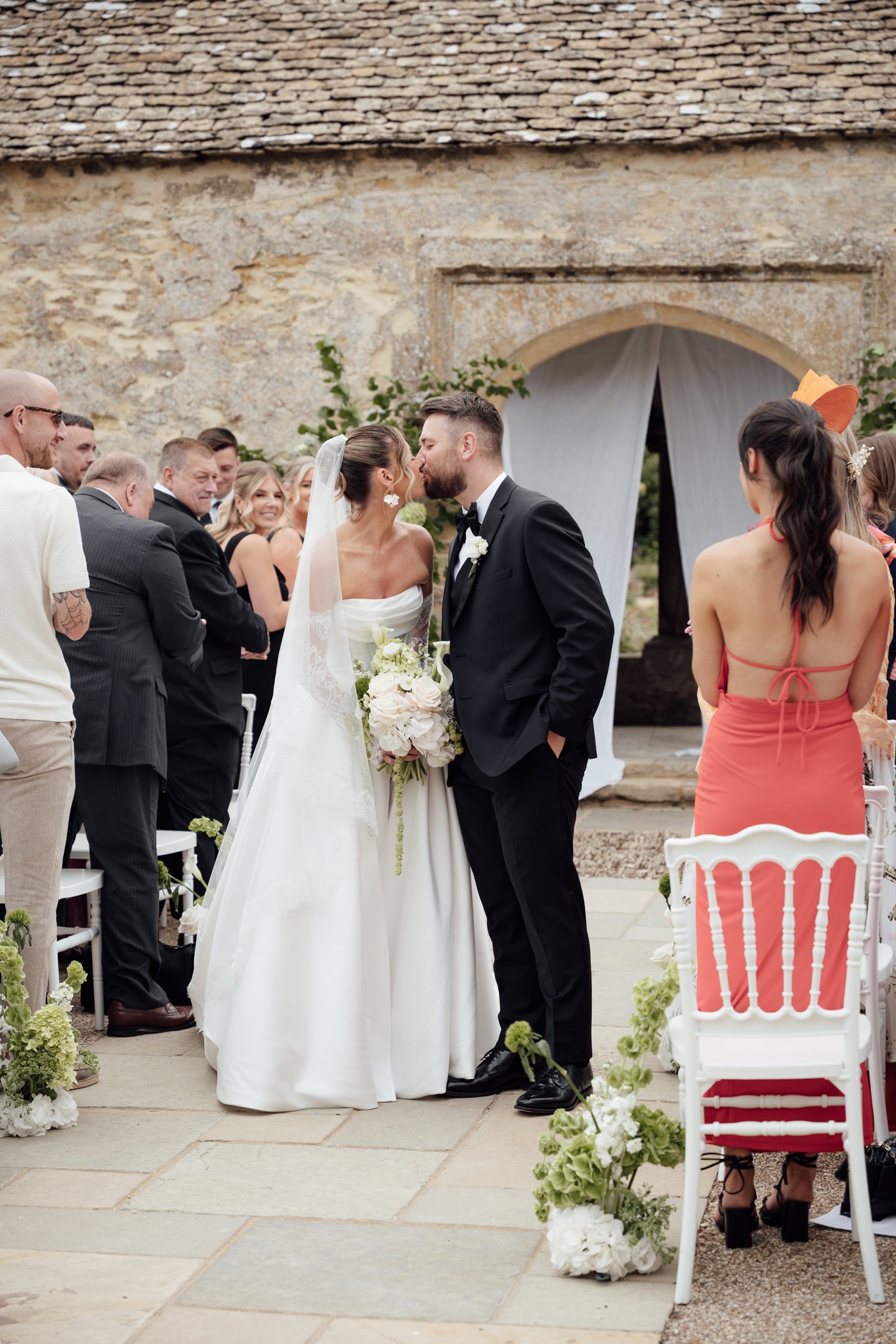 A bride and groom kiss during their outdoor wedding ceremony with guests and floral arrangements at Caswell House.