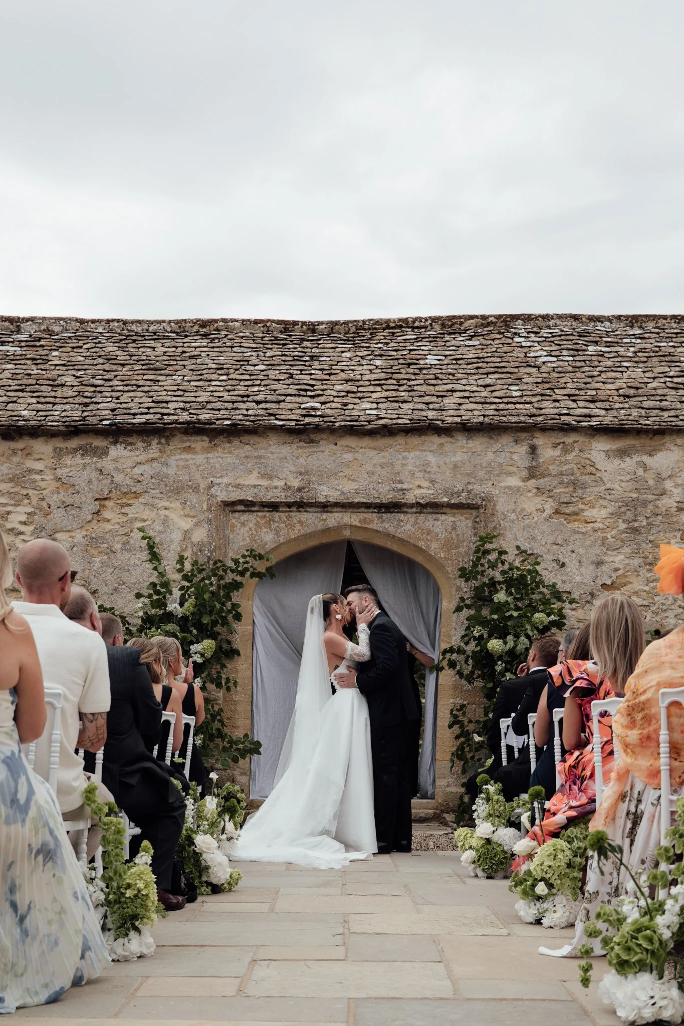 A bride and groom sharing their first kiss at Caswell House, standing beneath the stone archway as guests watch from the aisle.