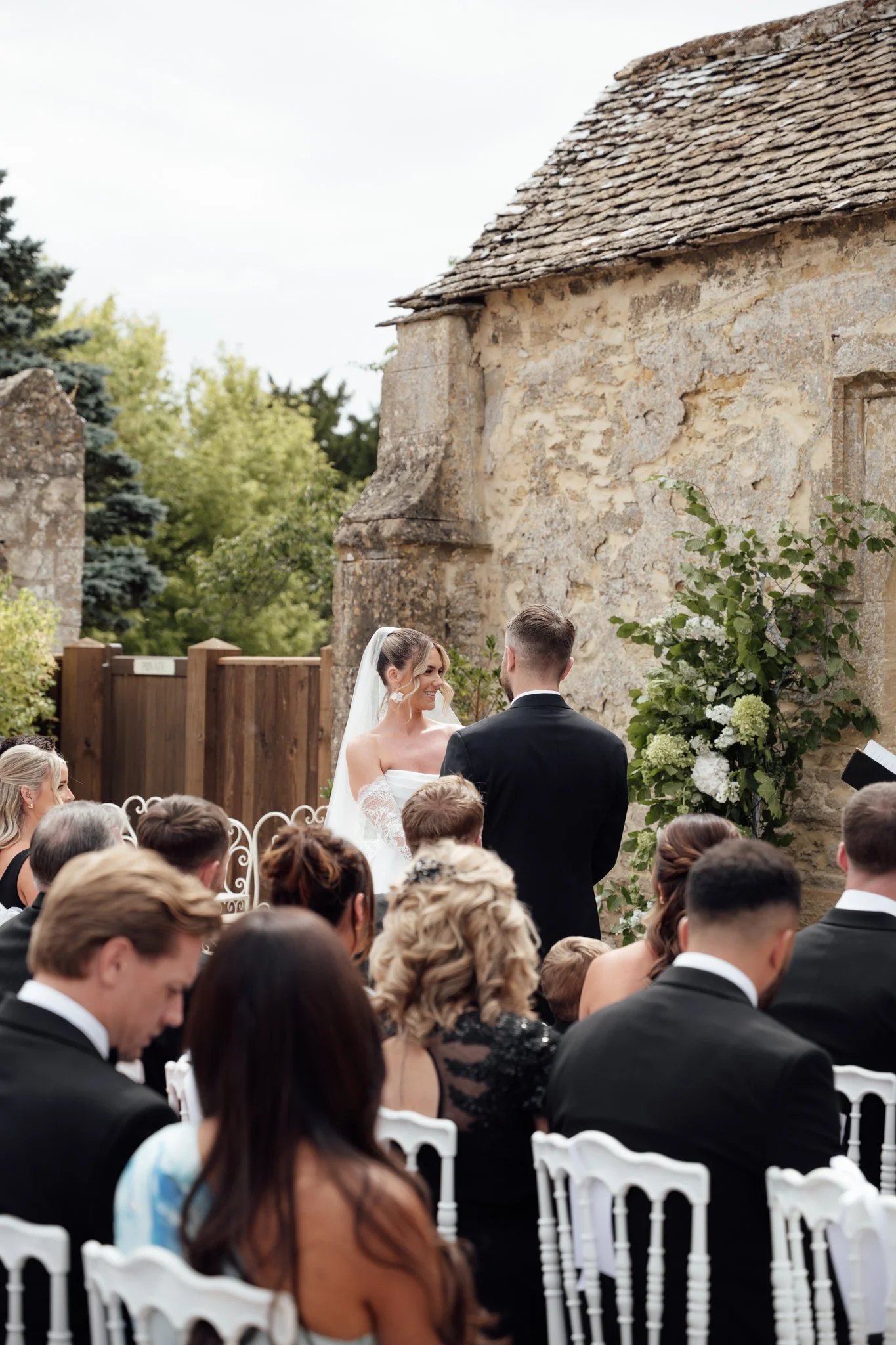 Guests seated at an outdoor ceremony at Caswell House as the bride and groom stand together at the front surrounded by the stone barn and greenery.