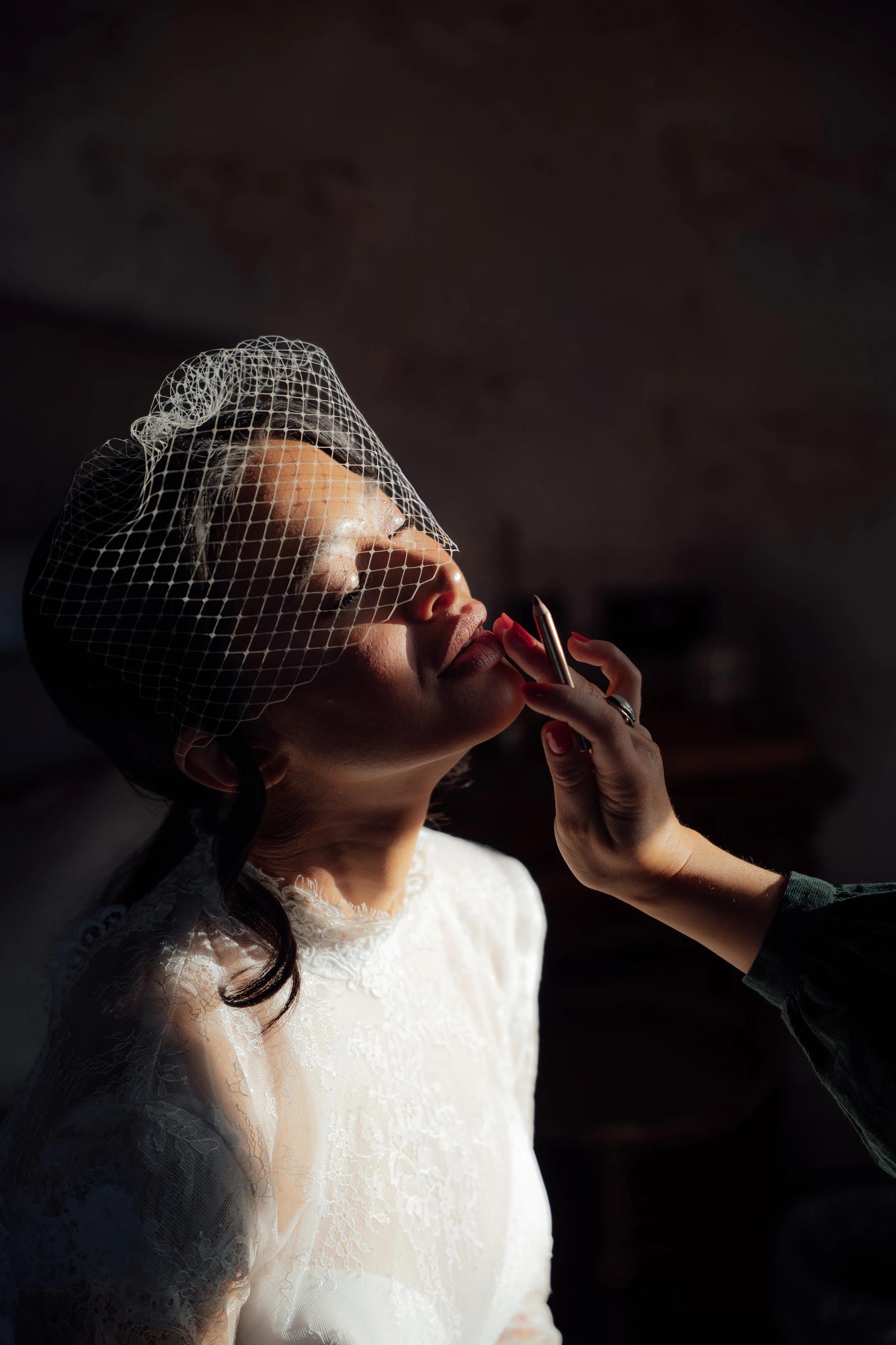 A dramatic getting-ready portrait at Cartmel Old Grammar, with the bride having her makeup applied in soft directional light while wearing a birdcage veil.