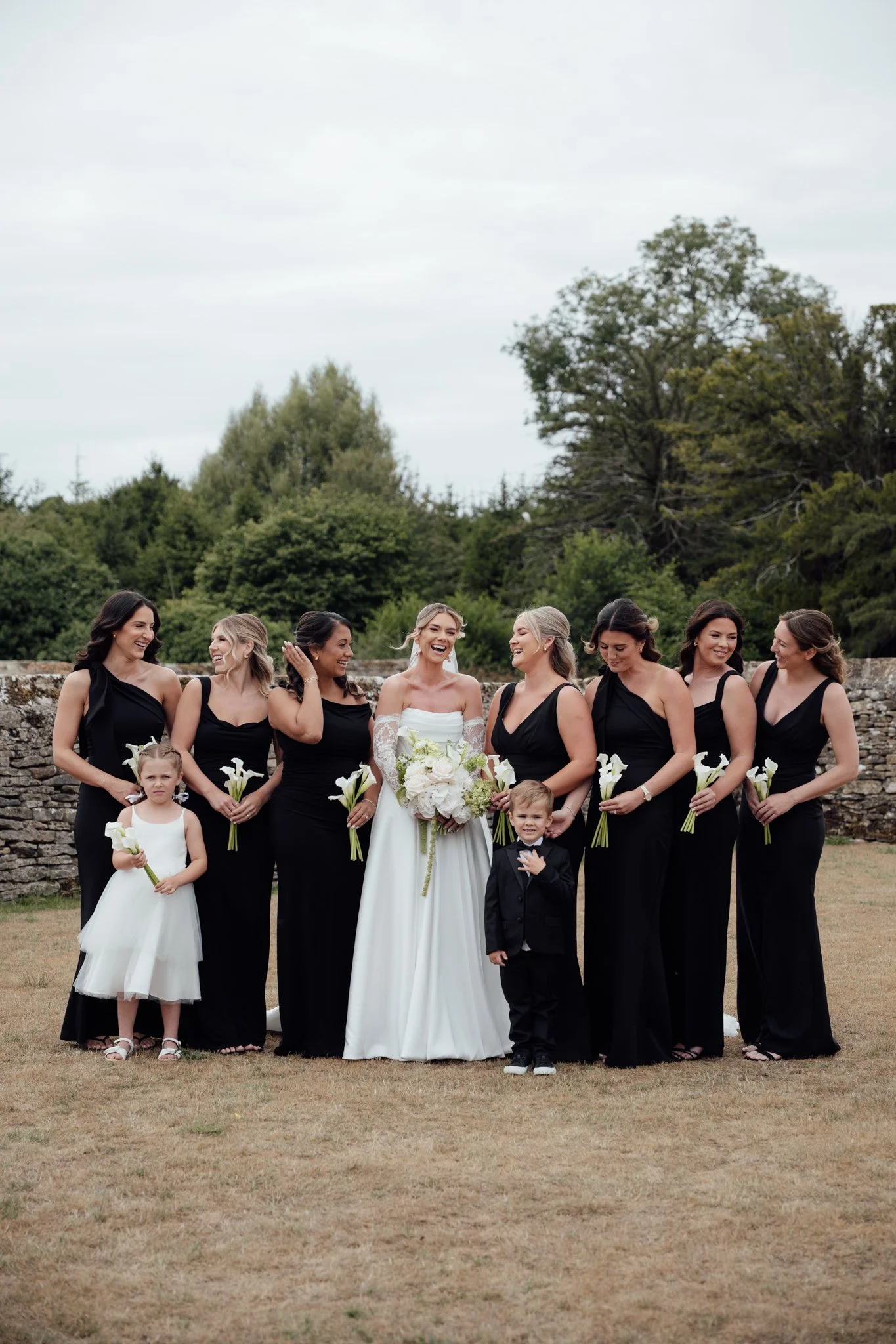 A bride stands with her bridesmaids holding bouquets, all dressed in black gowns, gathered together outdoors before the ceremony.