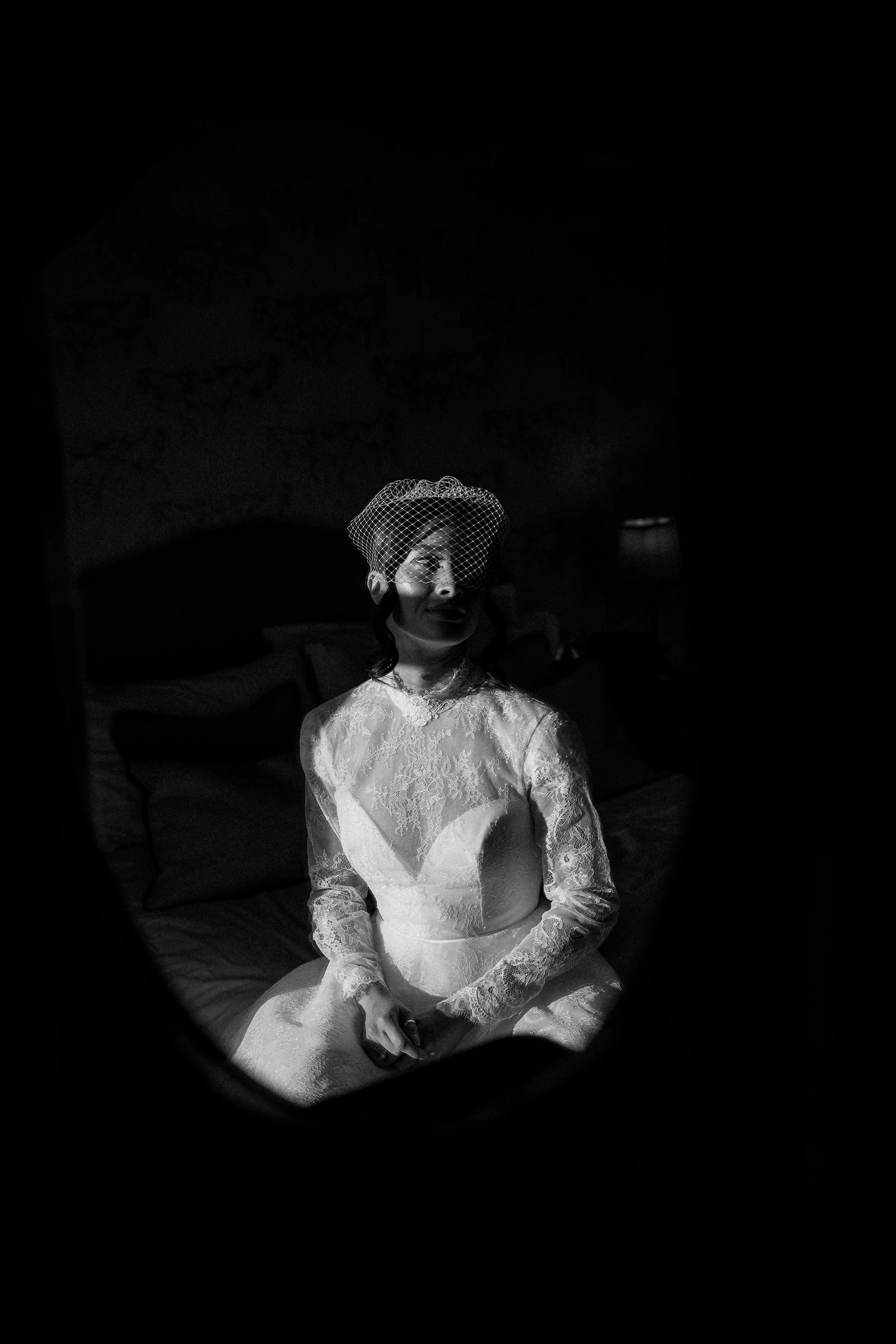 A bride sits in soft window light, her lace gown glowing gently in the shadows as she prepares for the day ahead, captured in an elegant and atmospheric moment.