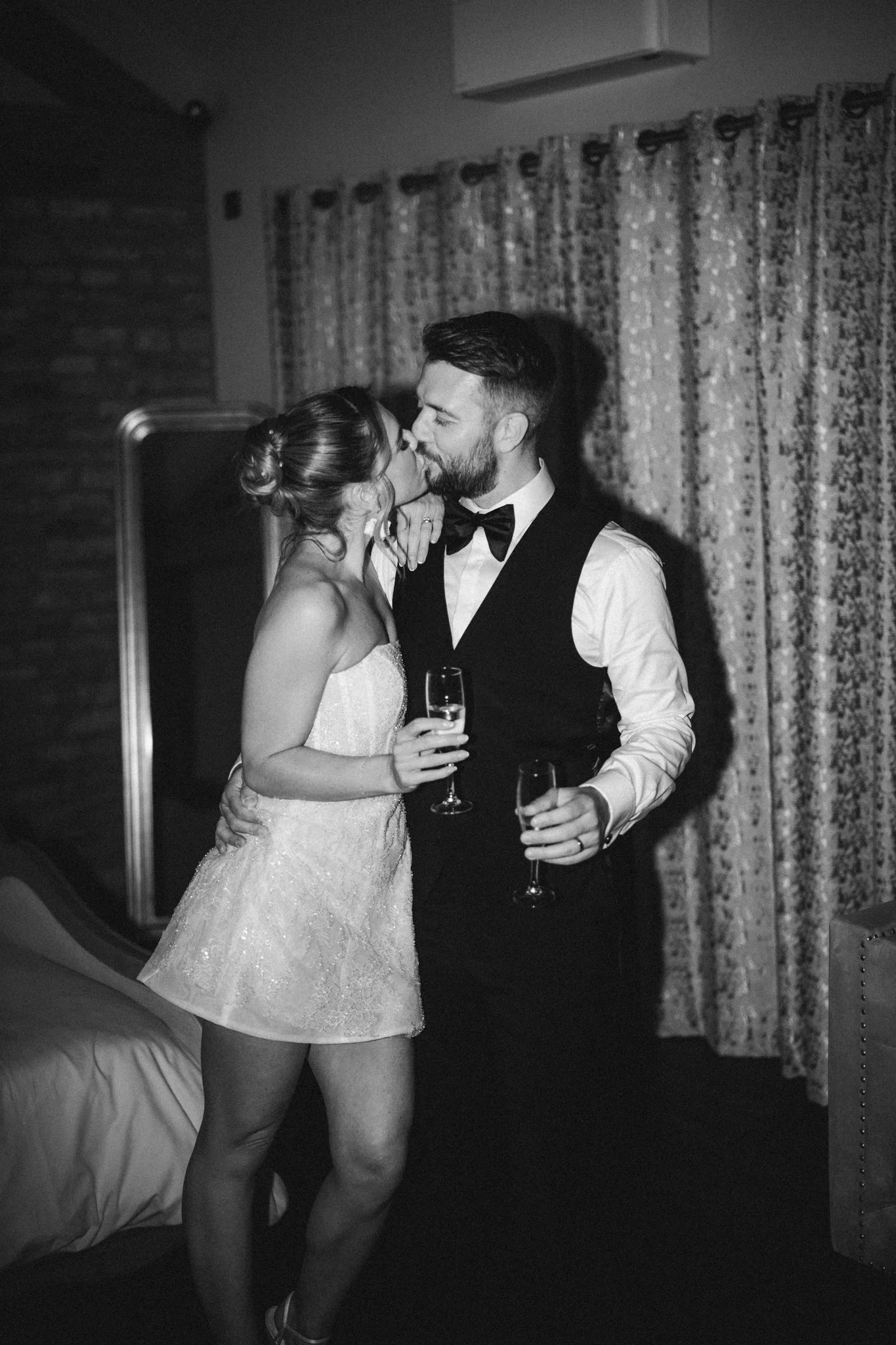 A black and white photo of a couple kissing at a celebration; the woman is in a strapless dress and holding a glass of champagne, the man is in a suit with a bow tie, also holding a glass of champagne.