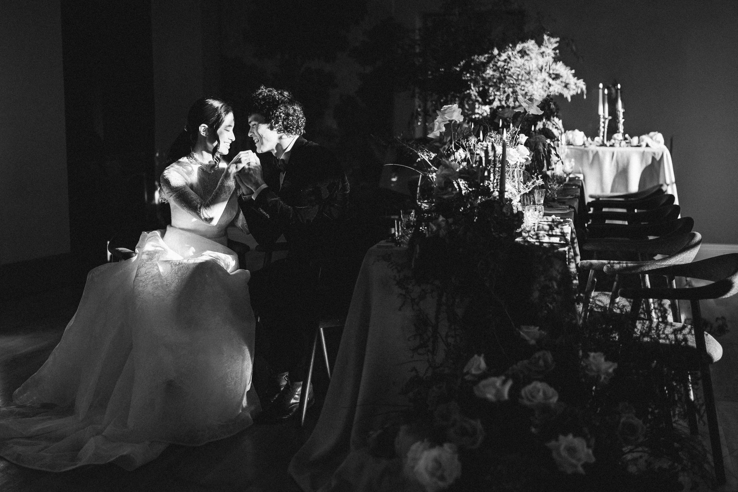 A bride and groom share an intimate moment beside a candlelit wedding table, surrounded by lush florals and dramatic shadows, at Cartmel Old Grammar House