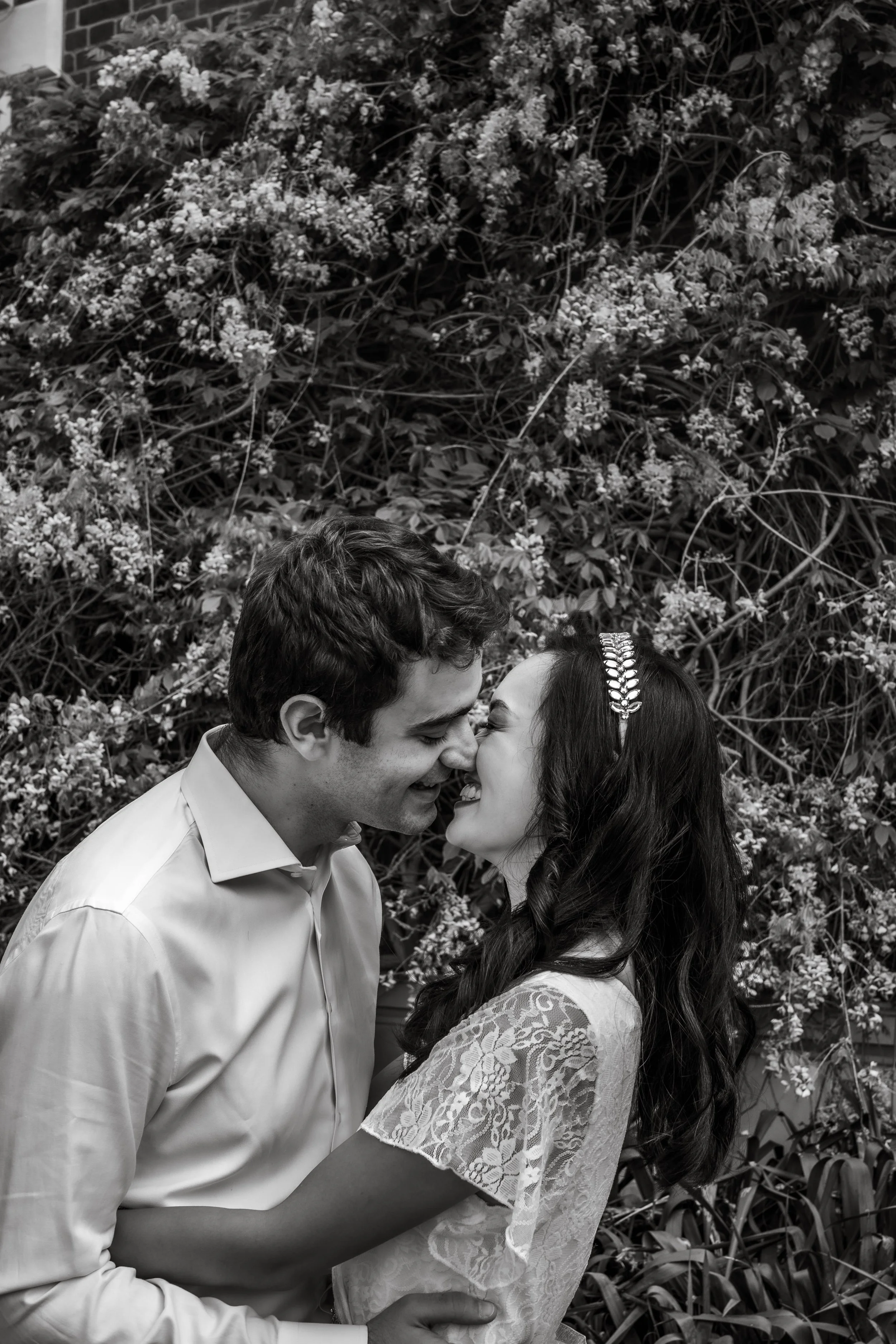 A man and woman sharing a kiss outdoors, surrounded by trees. The image is in black and white, capturing an intimate moment of romance between them. Both individuals are wearing clothing, with their faces expressing affection as they engage in this l
