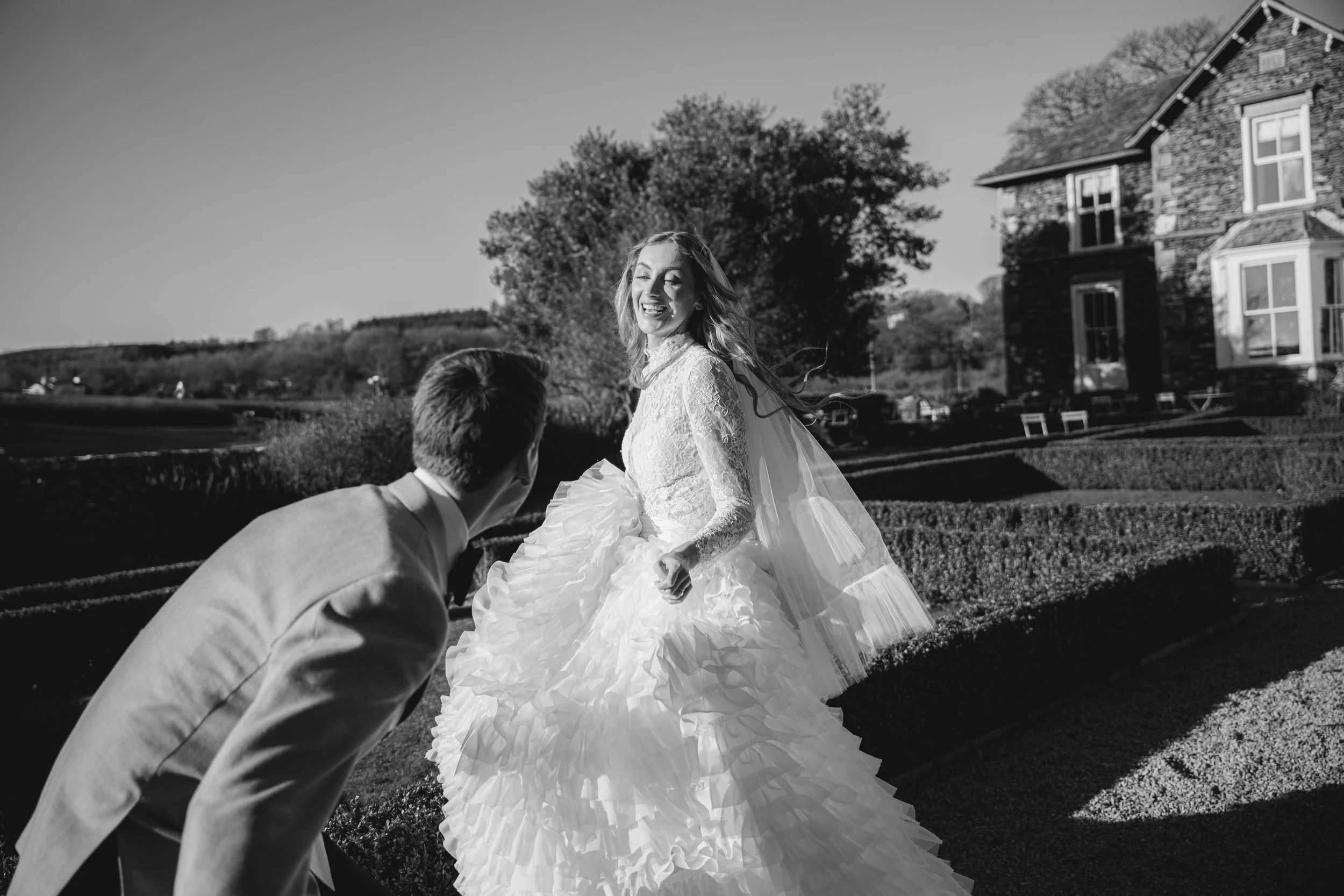 A joyful bride running through the gardens at Old Grammar House as her groom playfully reaches toward her, captured in soft evening light.