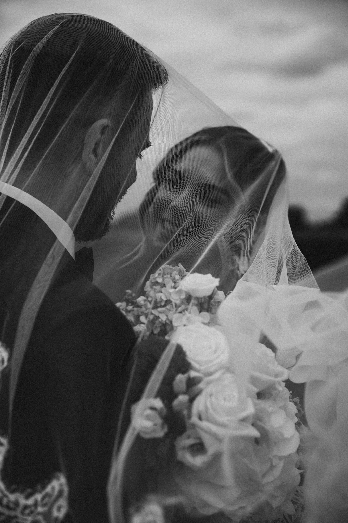 A black-and-white photo of a bride and groom close-up, smiling and looking at each other under a veil, with the bride holding a bouquet of flowers.