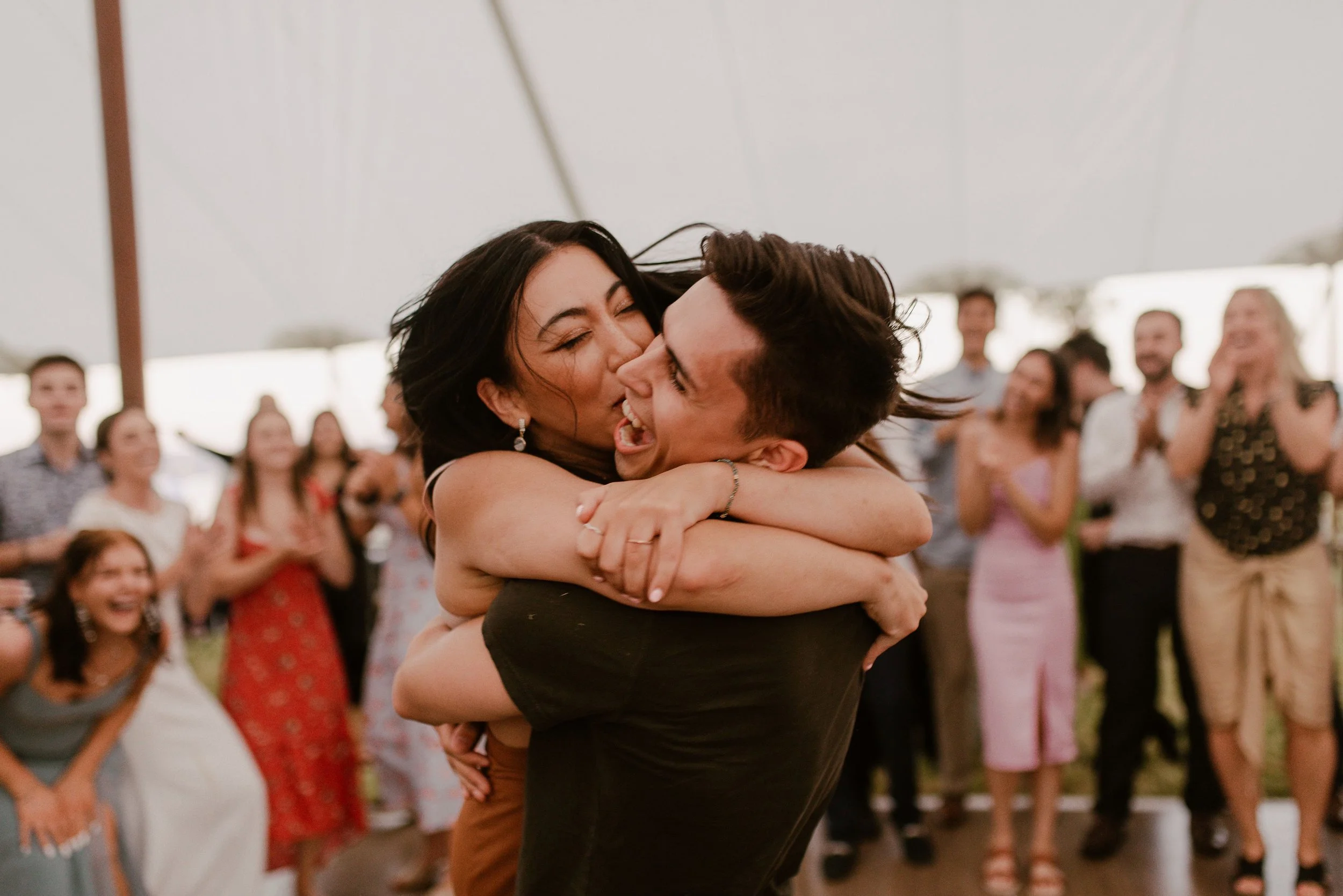 A couple is hugging at a social gathering, with a group of smiling people in the background under a large tent.