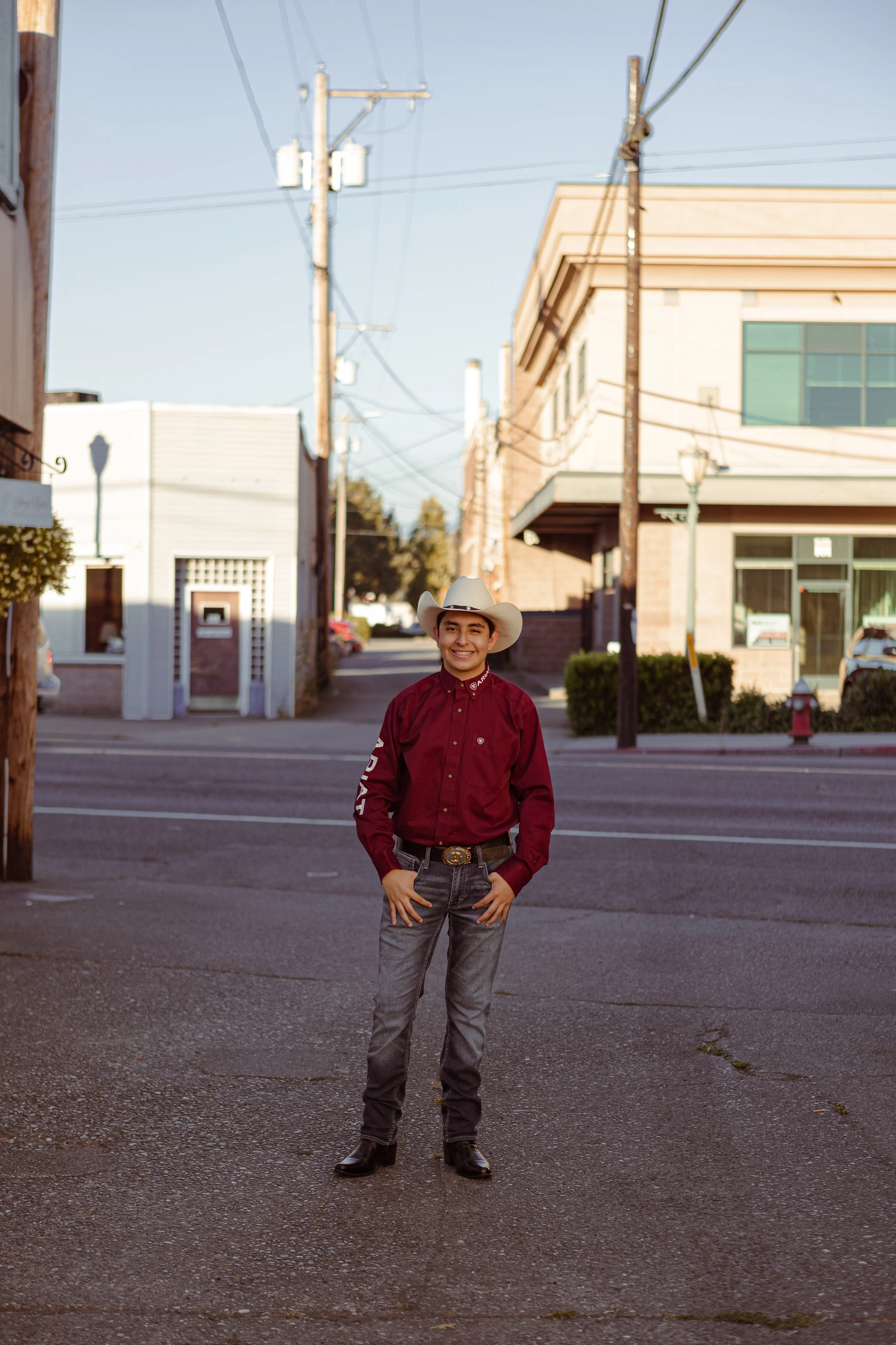 A young man dressed in a cowboy outfit with a white cowboy hat, maroon long-sleeve shirt, gray jeans, and black boots, standing on a city street with buildings, utility poles, and street lamps in the background during daylight.