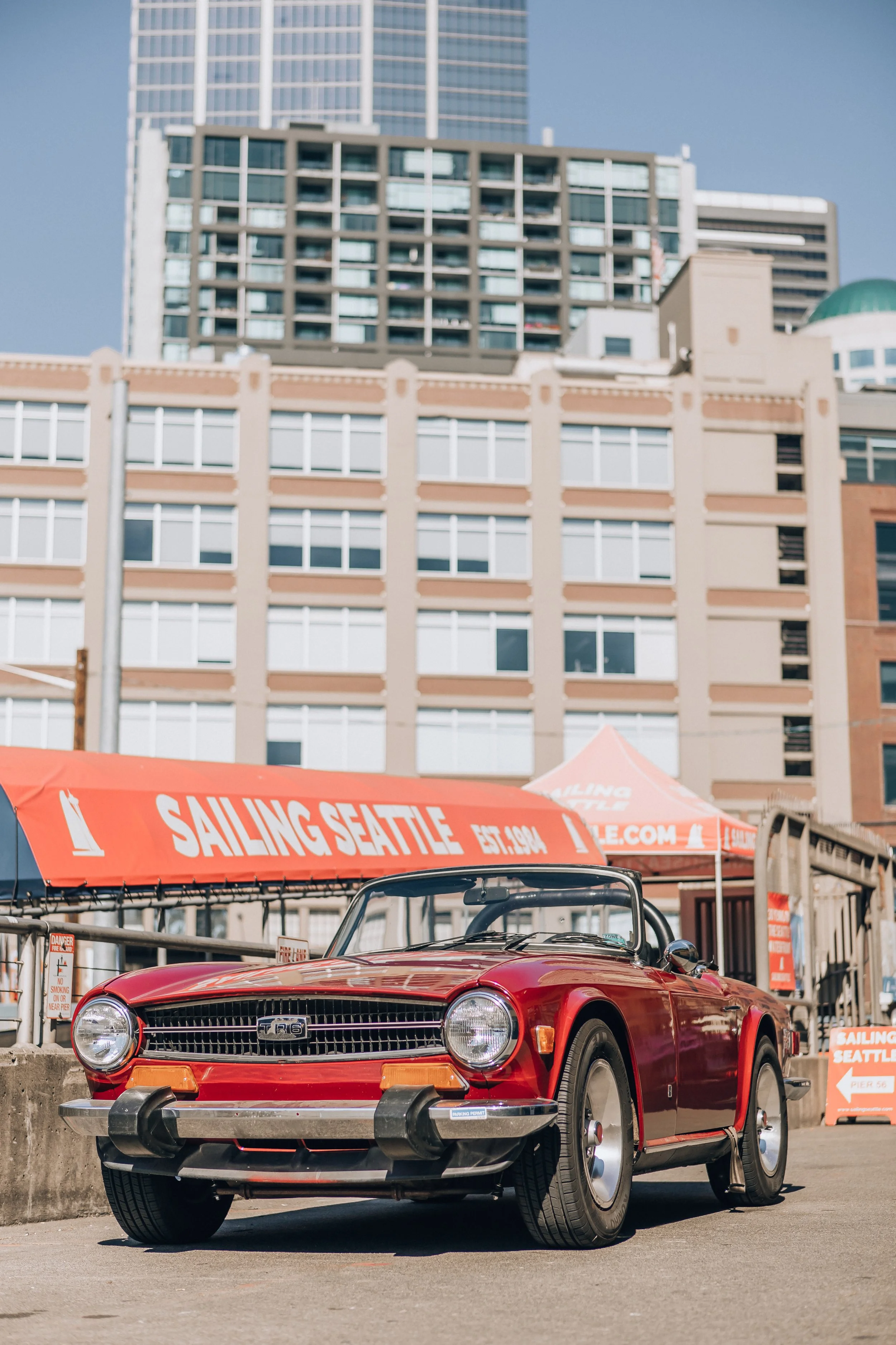 A red vintage Triumph TR6 convertible car parked in front of a concrete barrier with red sails advertising 'Sailing Seattle' at a waterfront event in an urban area. Tall buildings and tents are visible in the background.