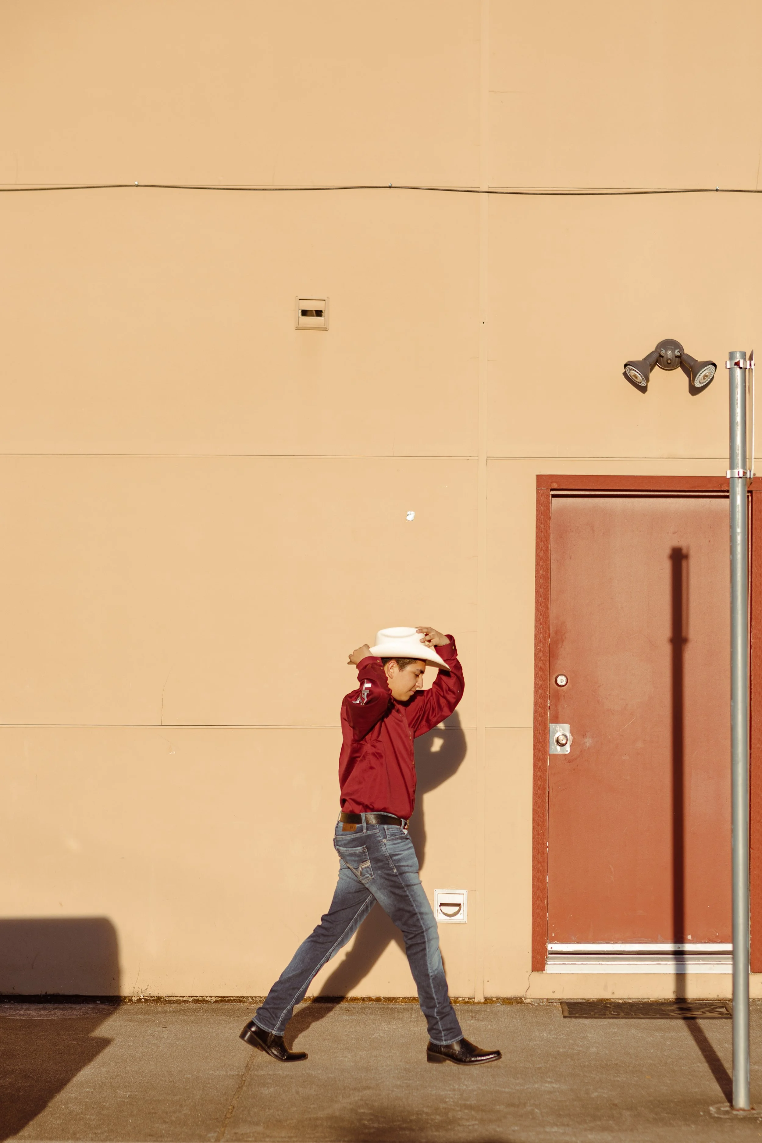 Young person in a red jacket and jeans walking along a beige wall while adjusting a white hat, with a door and two security cameras visible.