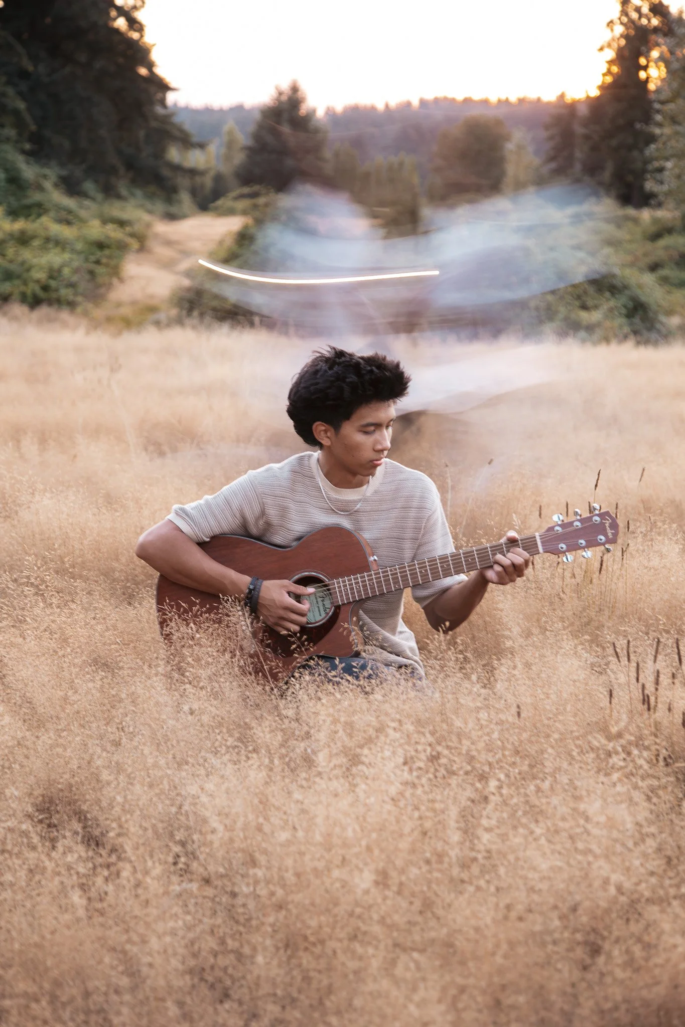 A young man with dark hair playing an acoustic guitar in a field of tall, golden grass during sunset.
