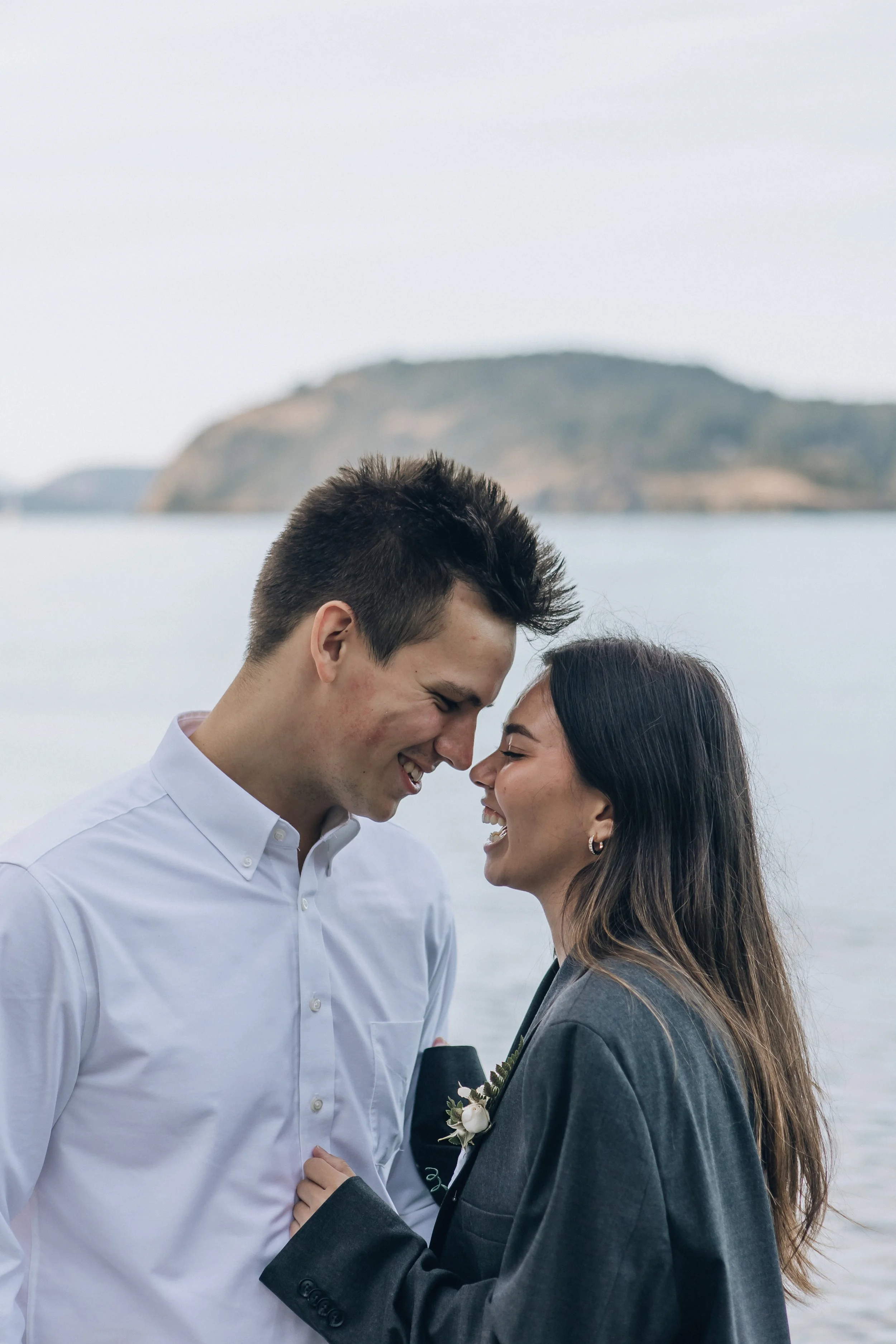 A couple smiling at each other outdoors near a body of water with a mountain in the background, dressed in formal attire.