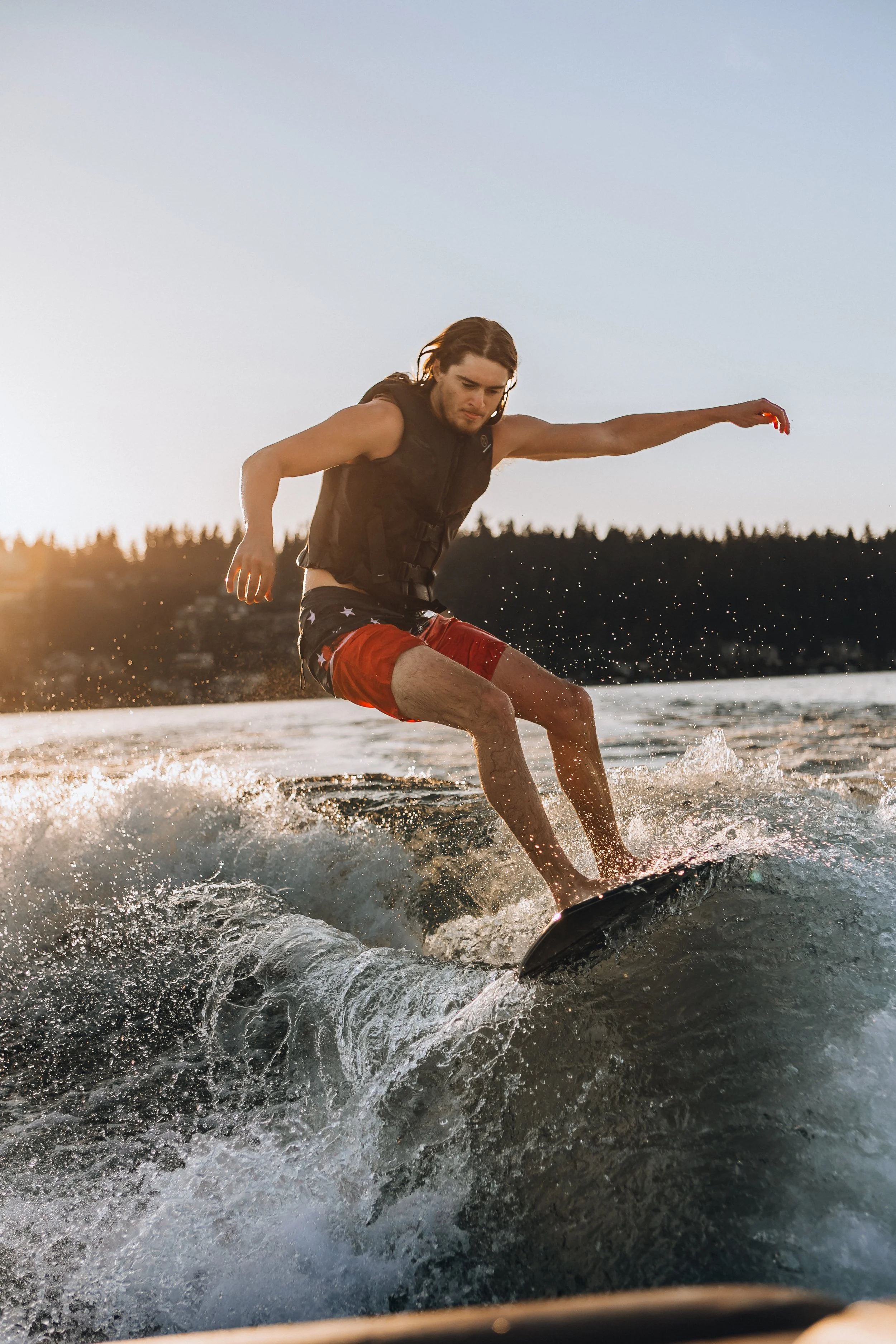A man with long brown hair stands on a wakeboard, surfing on a lake at sunset, wearing a black life jacket and red shorts with white stars.