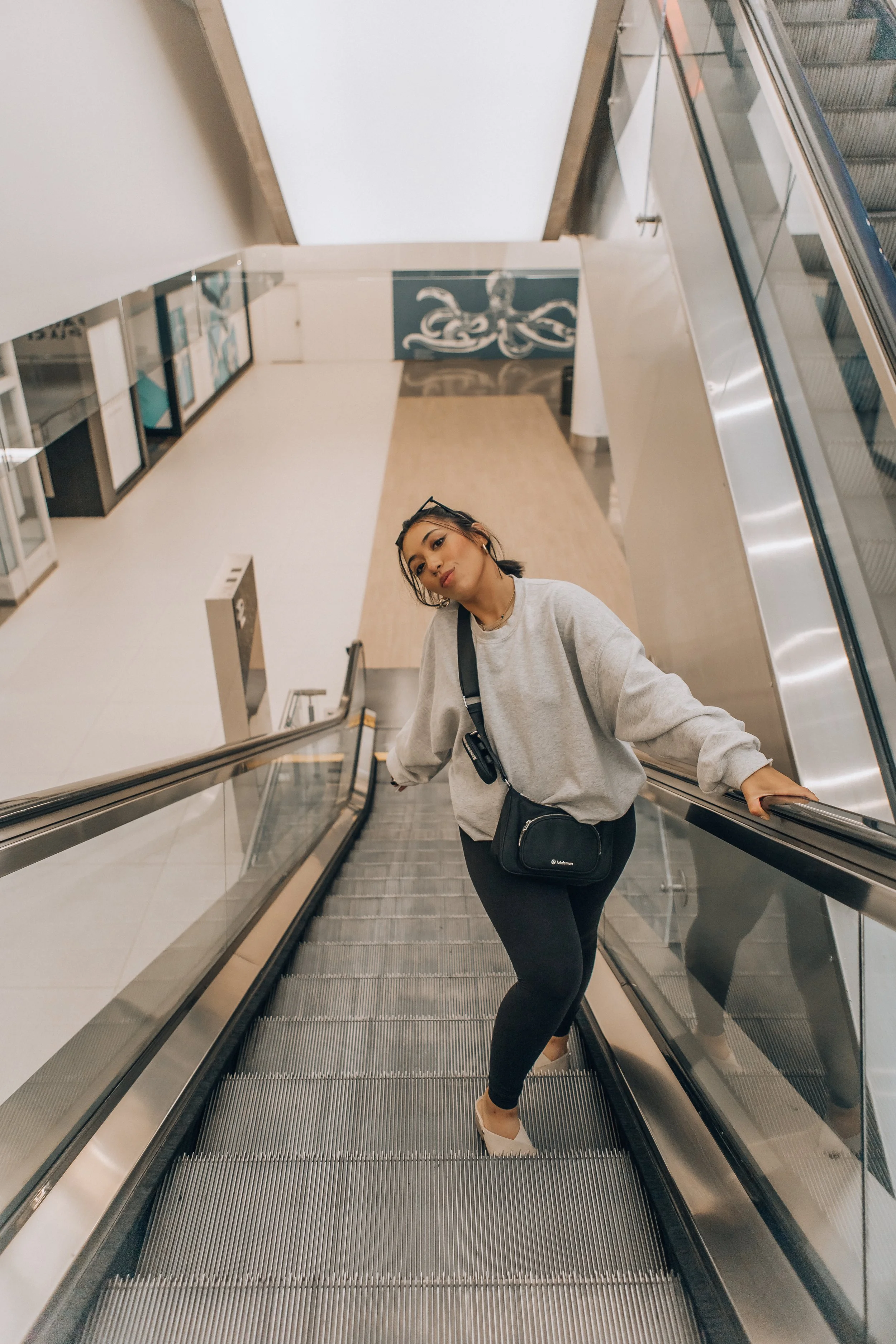 A young woman on an escalator in a modern indoor setting, wearing a gray sweatshirt, black pants, and white sneakers, holding onto the handrail, with sunglasses on her head and a black crossbody bag.