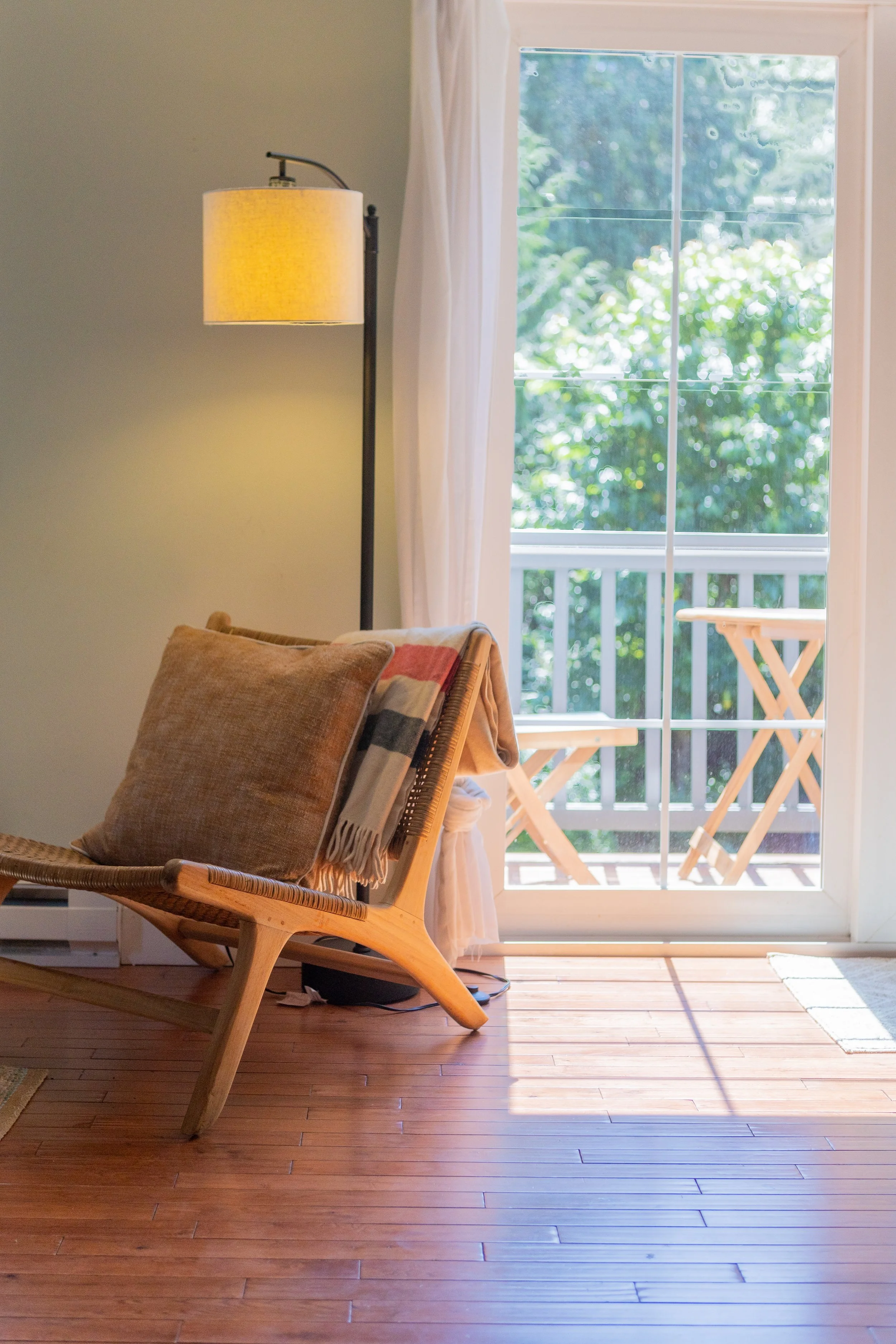 A wooden chair with a brown and beige pillow and throw blanket, a floor lamp with a yellow shade, and a sliding glass door leading to a balcony with greenery outside.