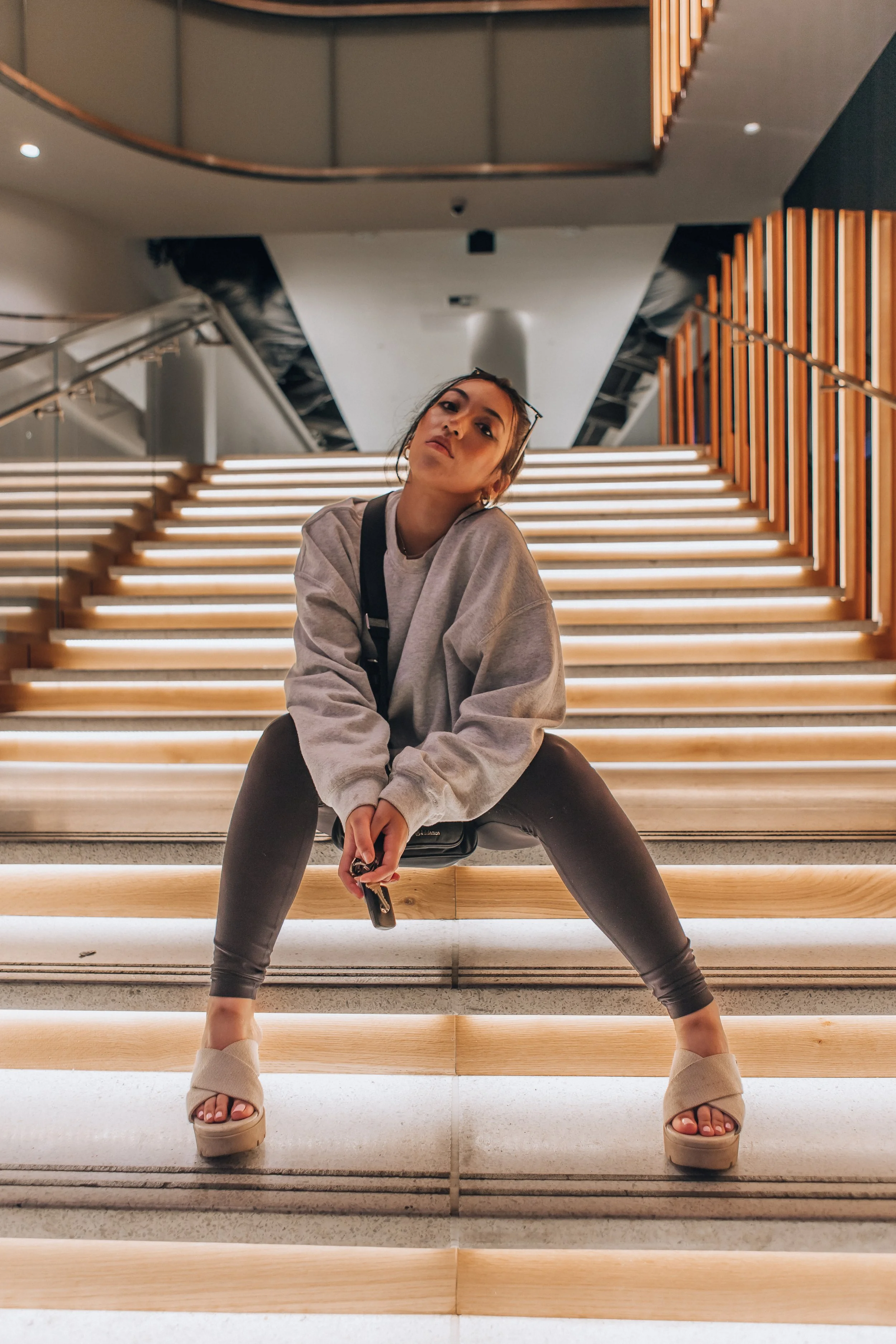 A woman sitting on the steps of a modern interior staircase with wooden steps and glass railing, posing with legs apart and holding sunglasses, wearing a gray oversized sweatshirt, dark leggings, and beige platform sandals.