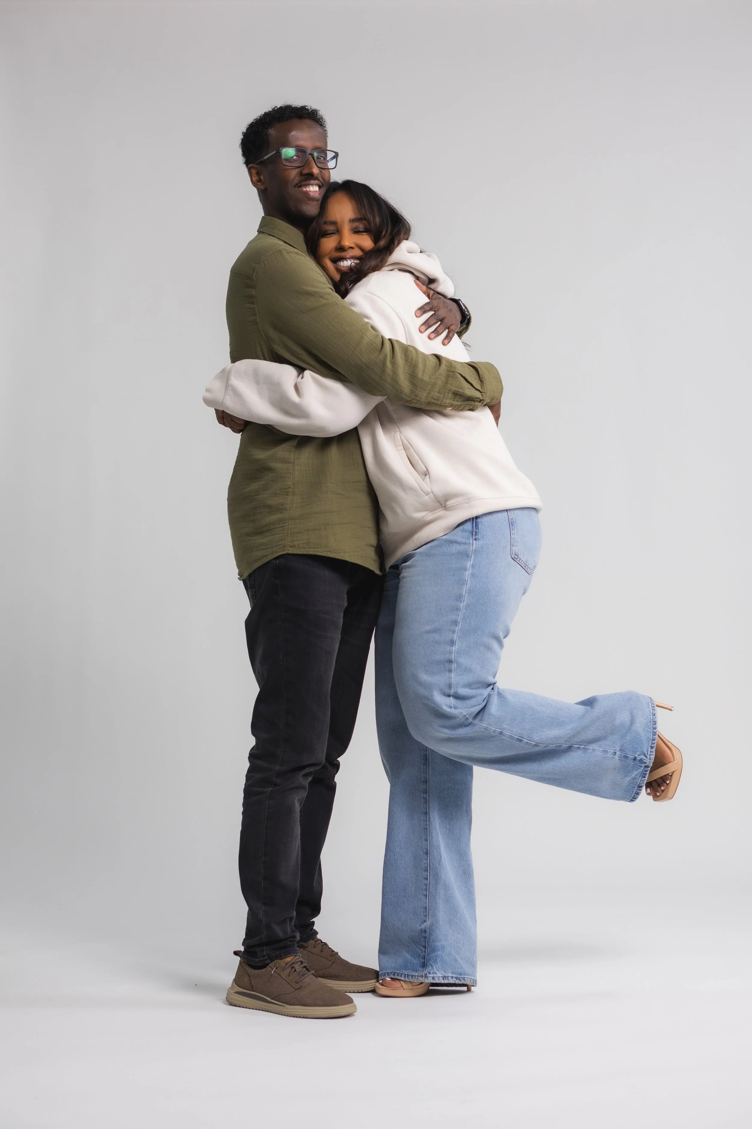 A man and woman hugging each other, with the woman lifting her leg and smiling, against a plain light gray background.