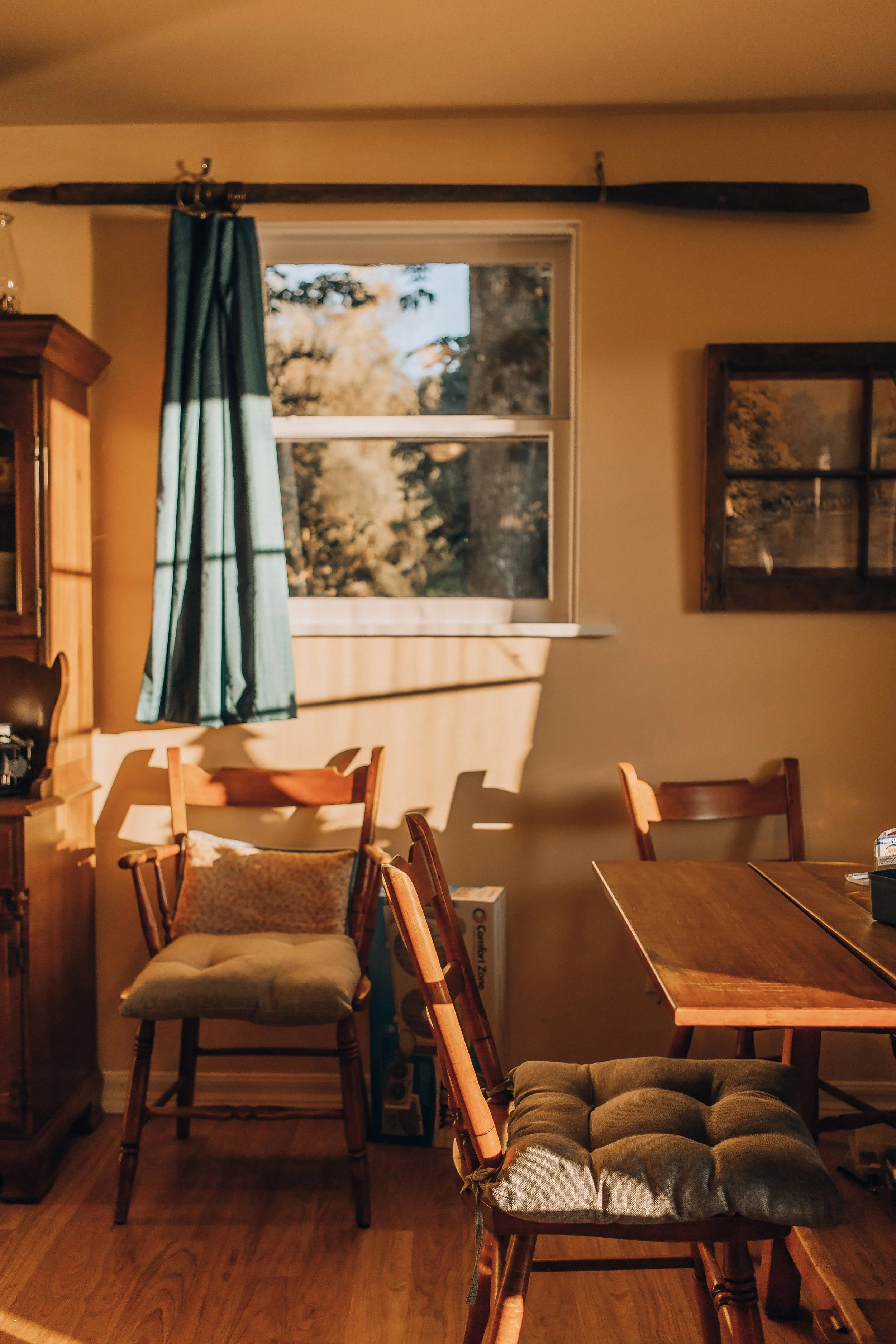Interior of a cozy dining room with wooden furniture, a window with a dark curtain, and sunlight casting shadows on the walls.