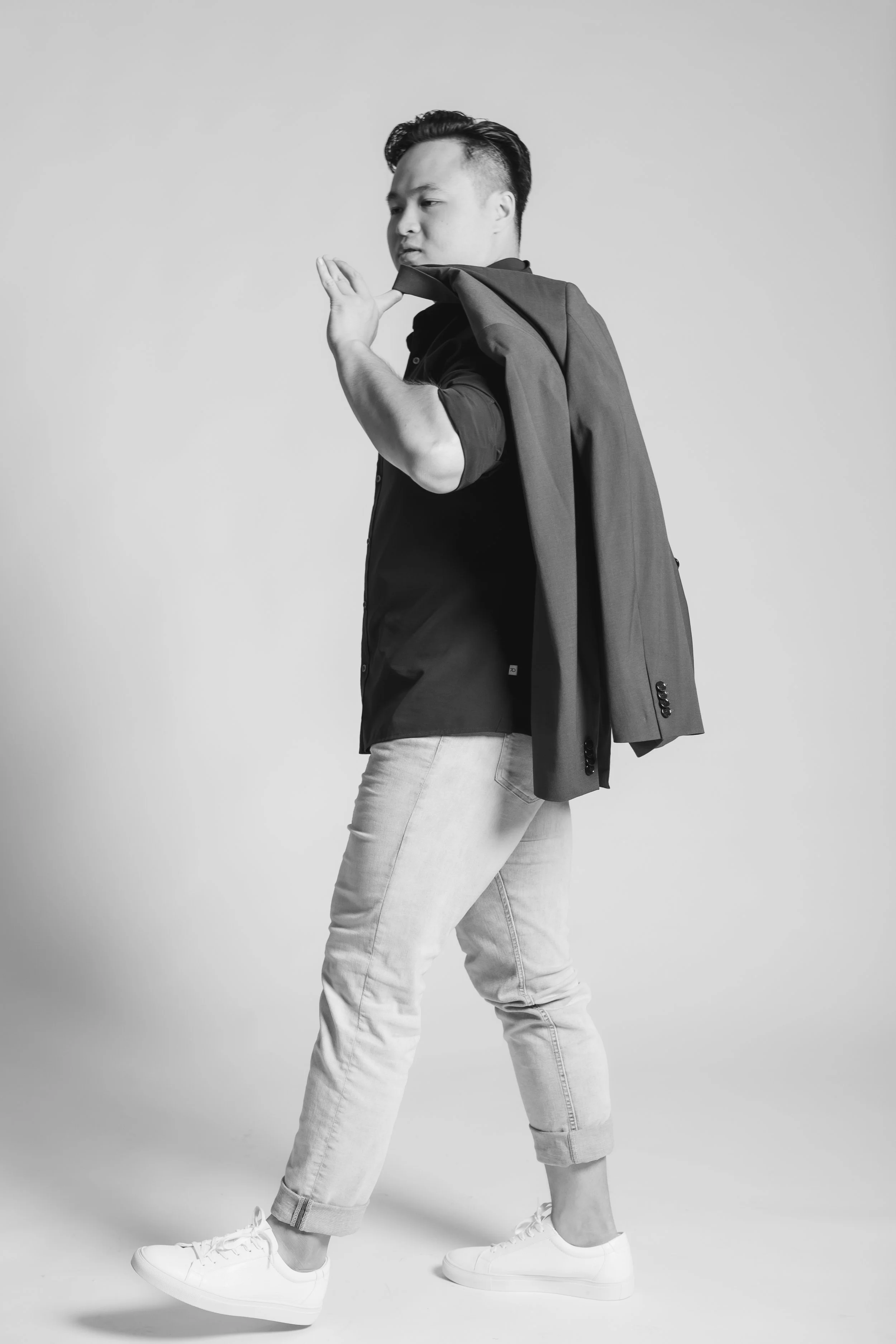 Black and white photo of a young man with styled hair, wearing a dark shirt and light jeans, holding a blazer over his shoulder, standing against a plain background.