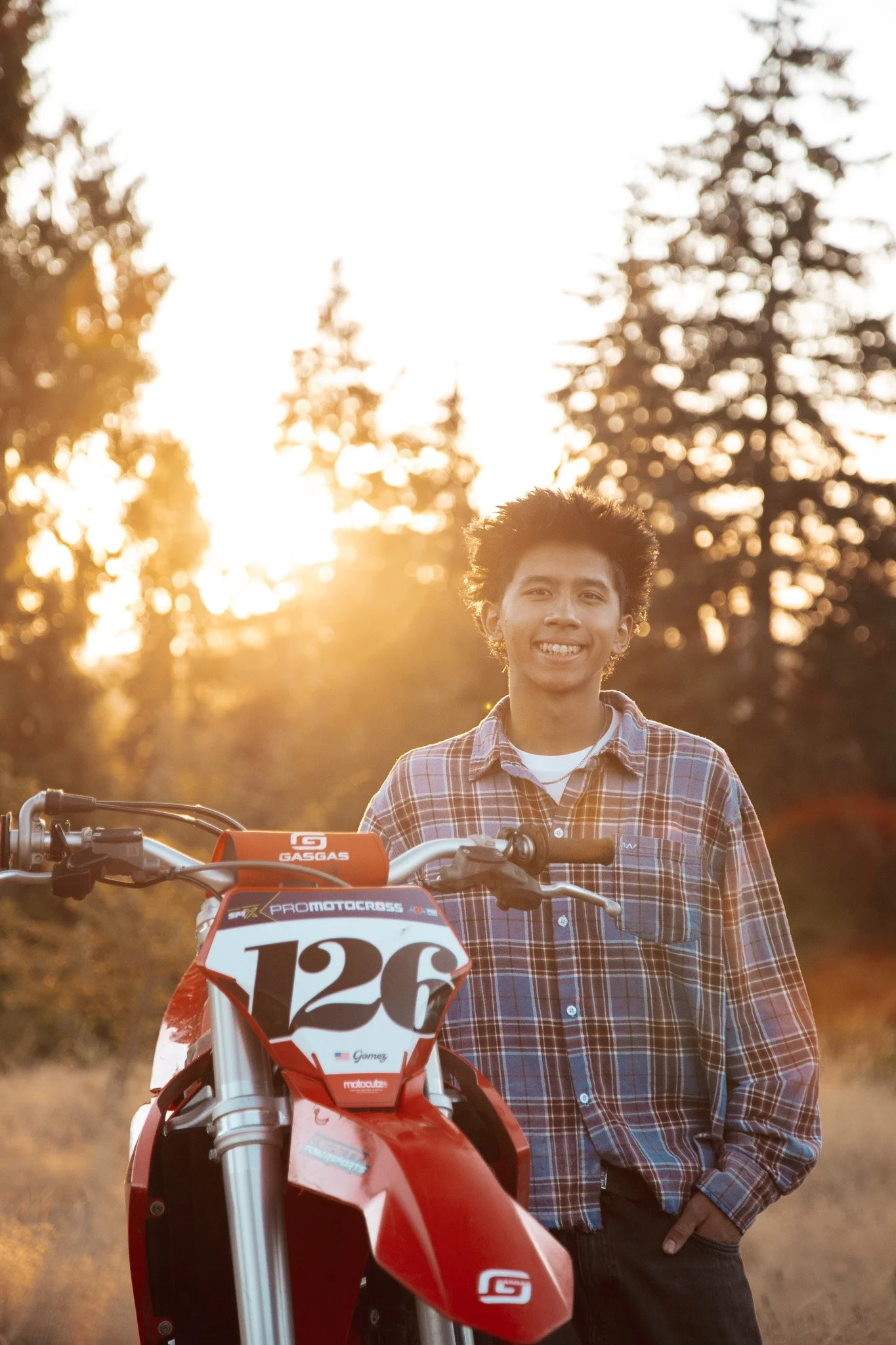 A young man in a plaid shirt standing outdoors at sunset, smiling, with a red dirt bike in front of him, trees in the background.