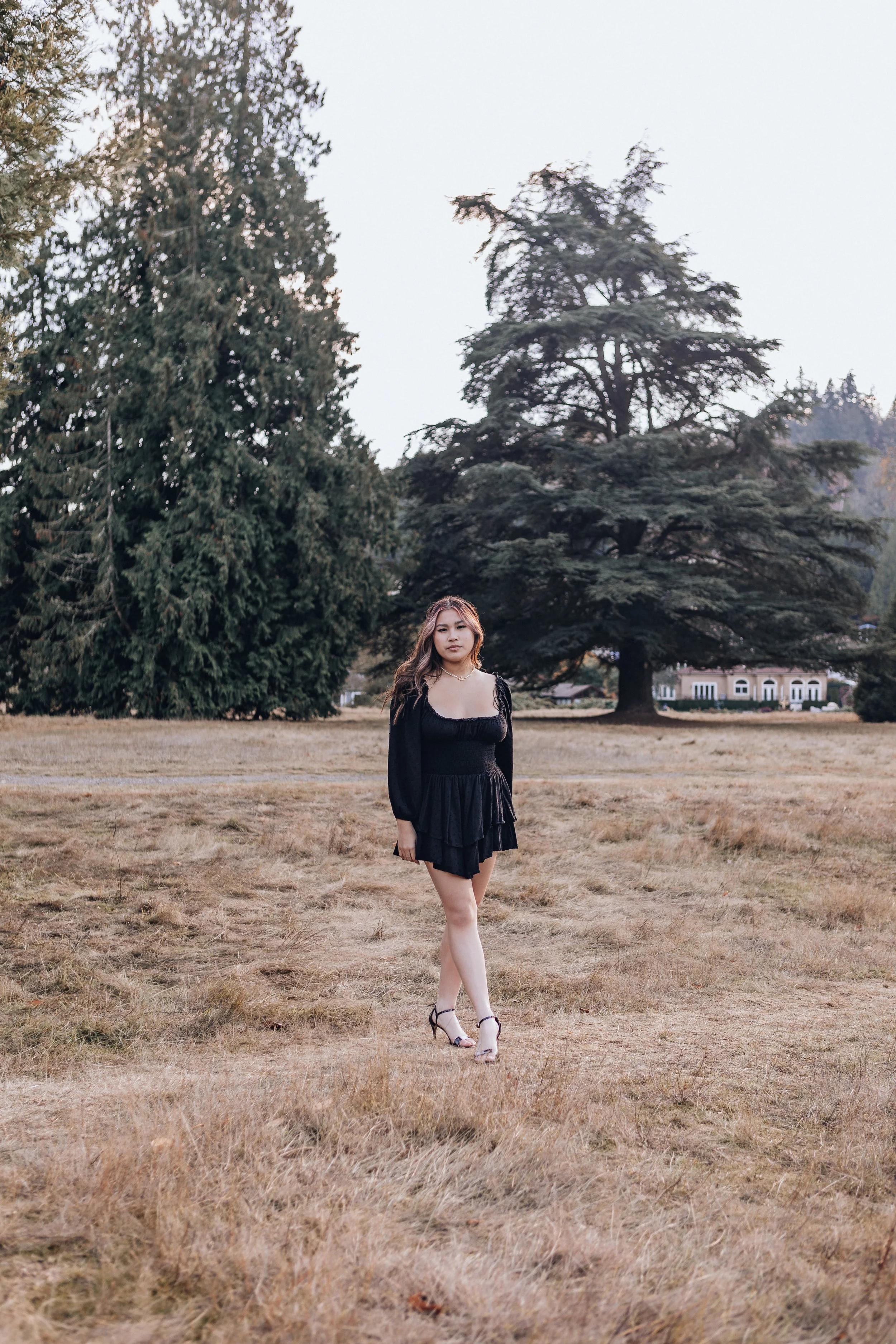 A young woman in a black dress and high heels walking through a grassy field with large trees in the background.