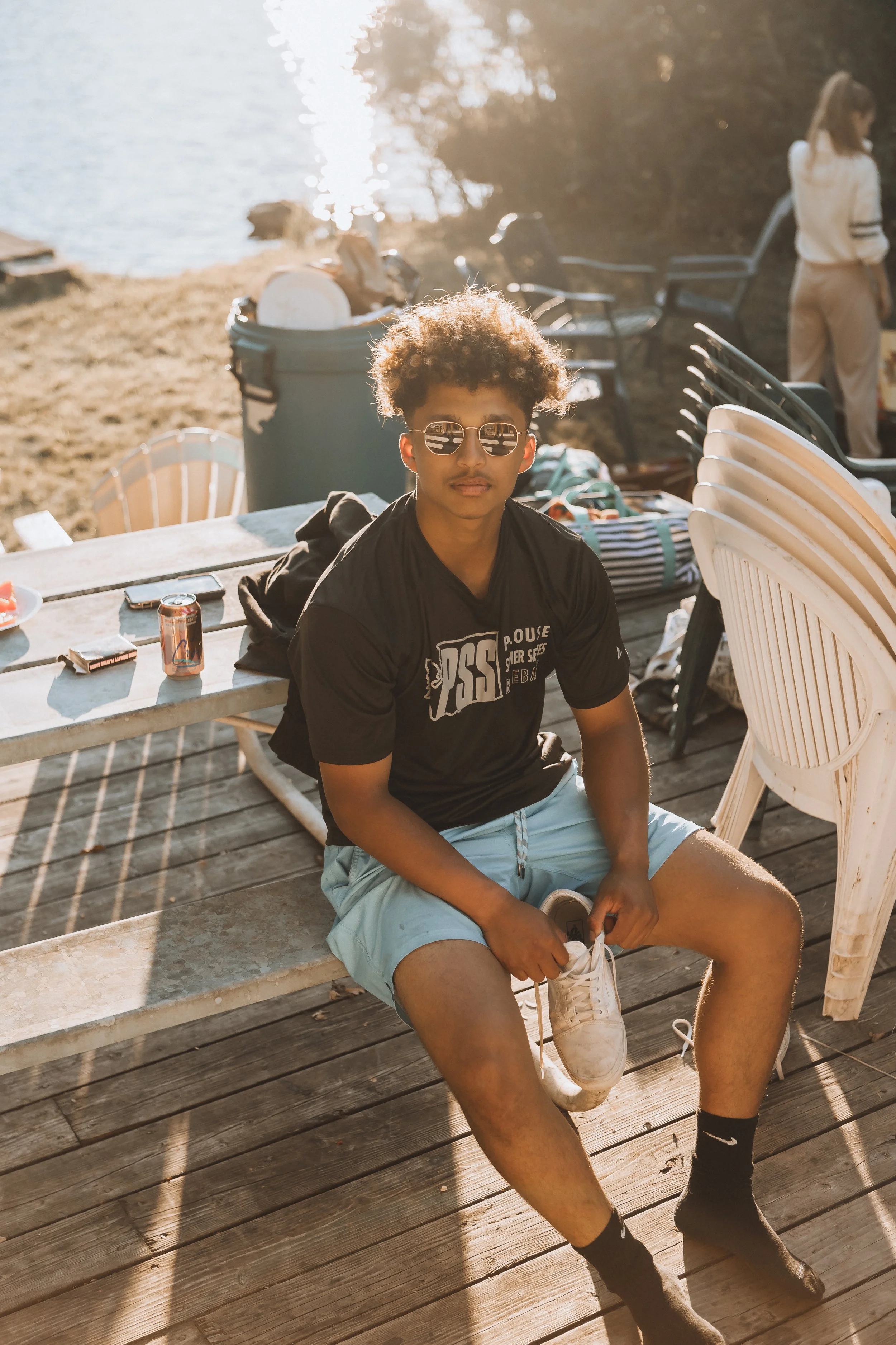 Young man with curly hair and sunglasses sitting on a wooden dock near a body of water, wearing a black T-shirt and light blue shorts, surrounded by chairs, containers, and other outdoor items, with sunlight reflecting off the water.