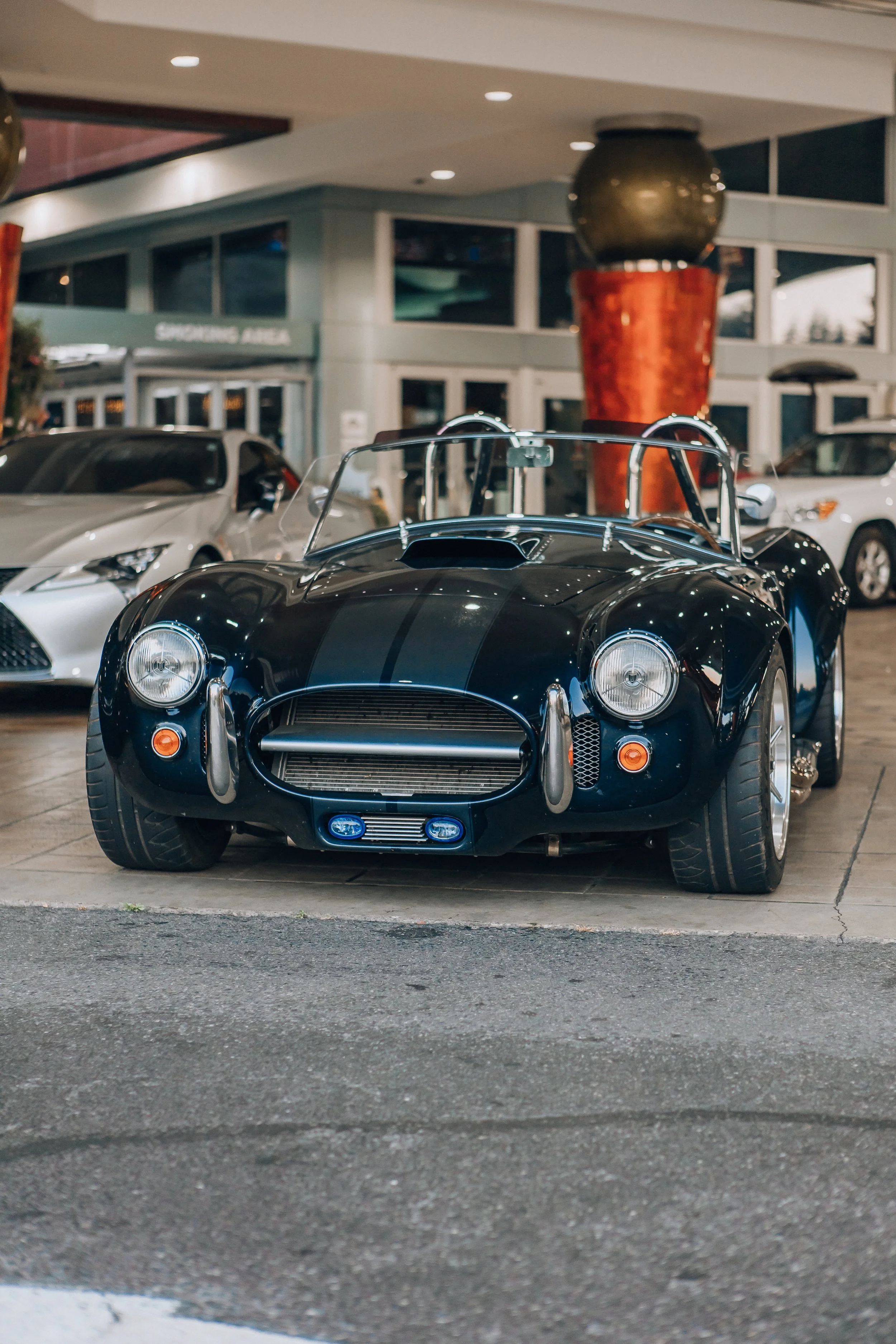 A black vintage sports car parked in front of a shopping mall, with other modern cars nearby and a large decorative sculpture behind it.
