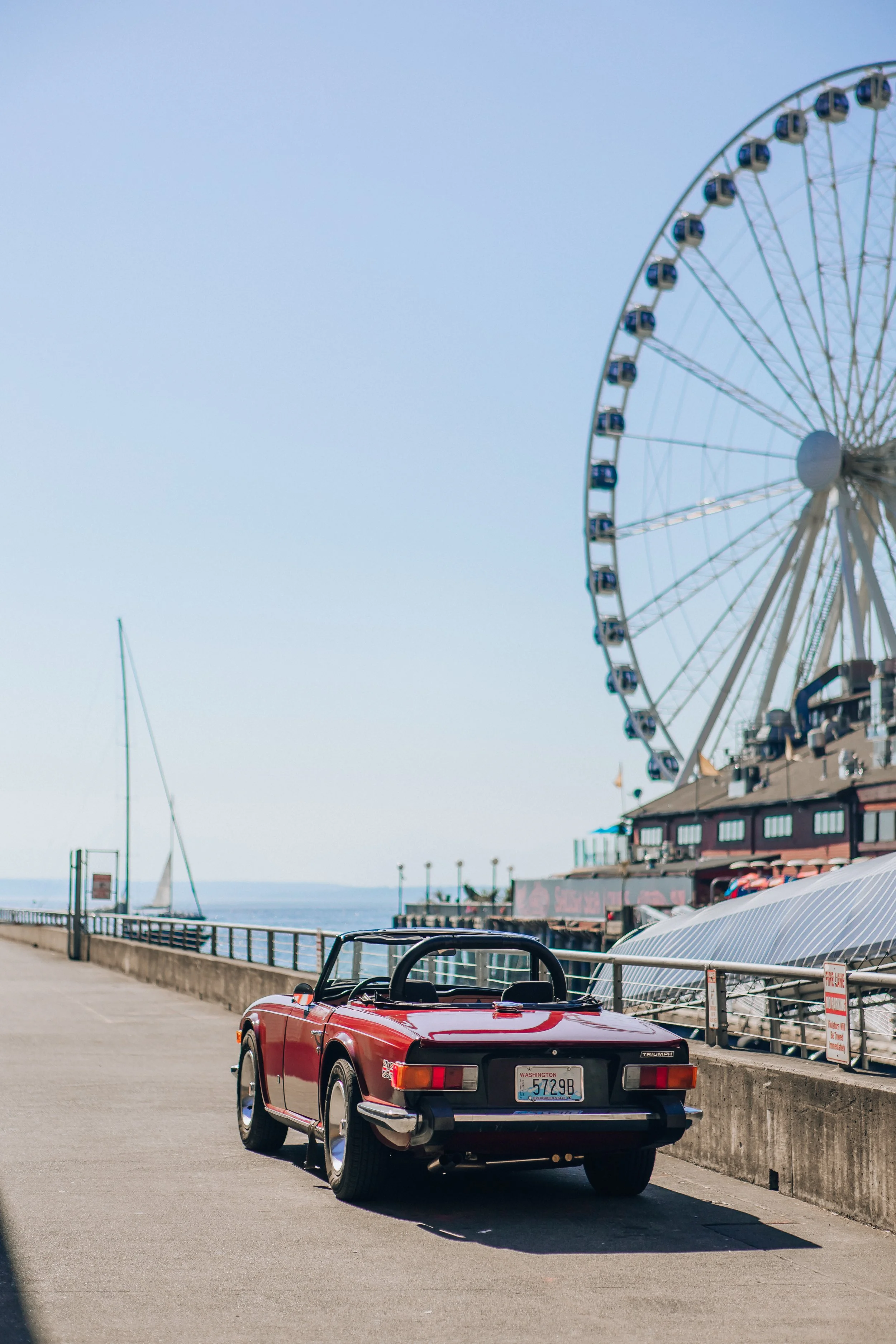 A red vintage convertible sports car parked along a waterfront promenade with a Ferris wheel and sailboats in the background on a sunny day.