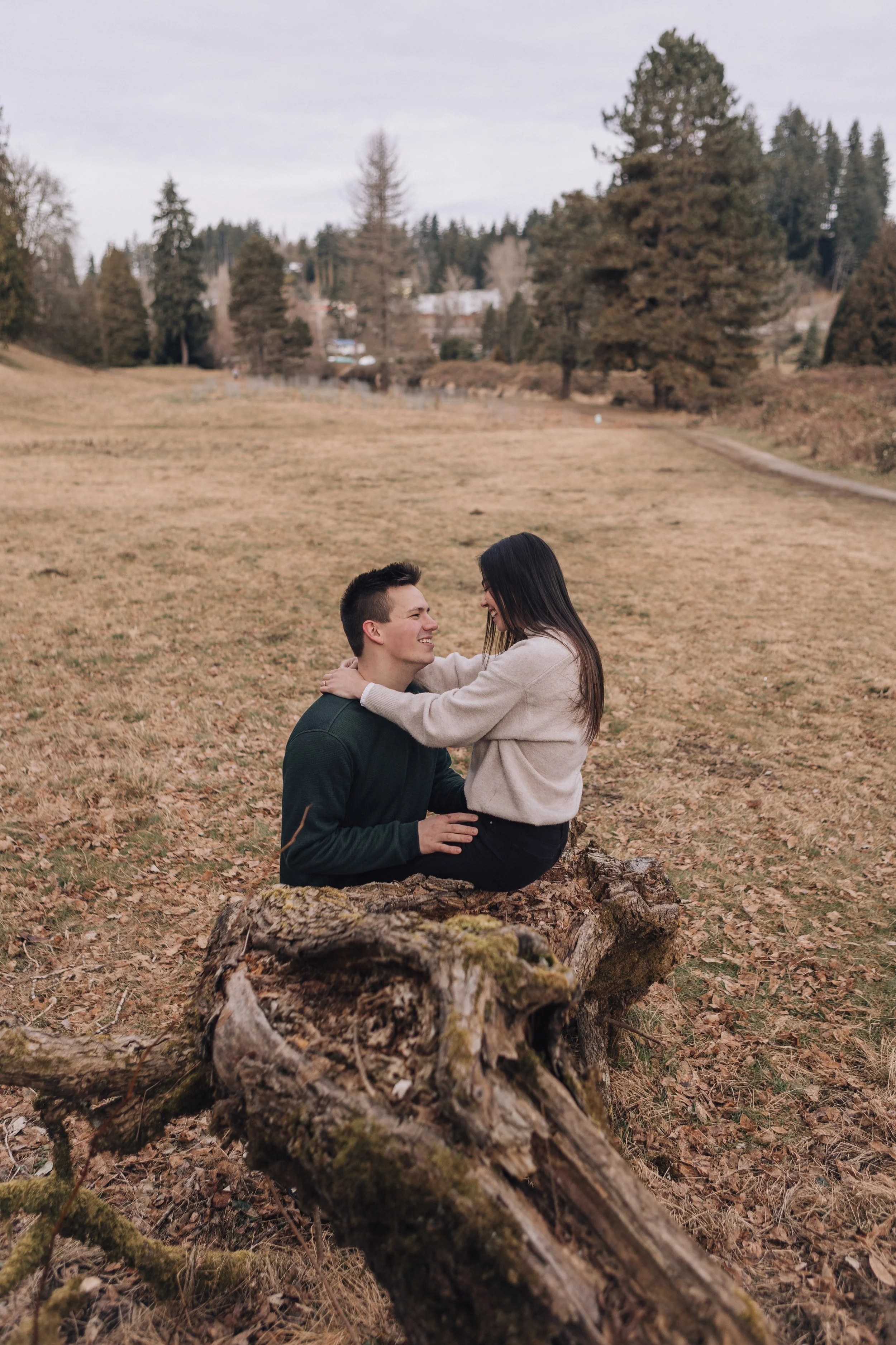 A young couple sitting on a fallen log in a park with trees and grass, smiling and embracing each other during daytime.
