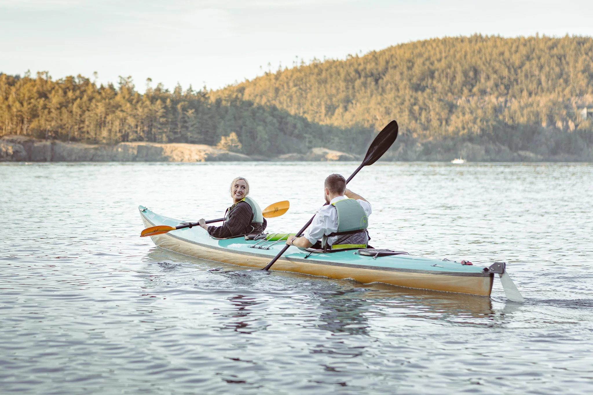 Two kayakers paddling on calm water beside a forested shoreline.