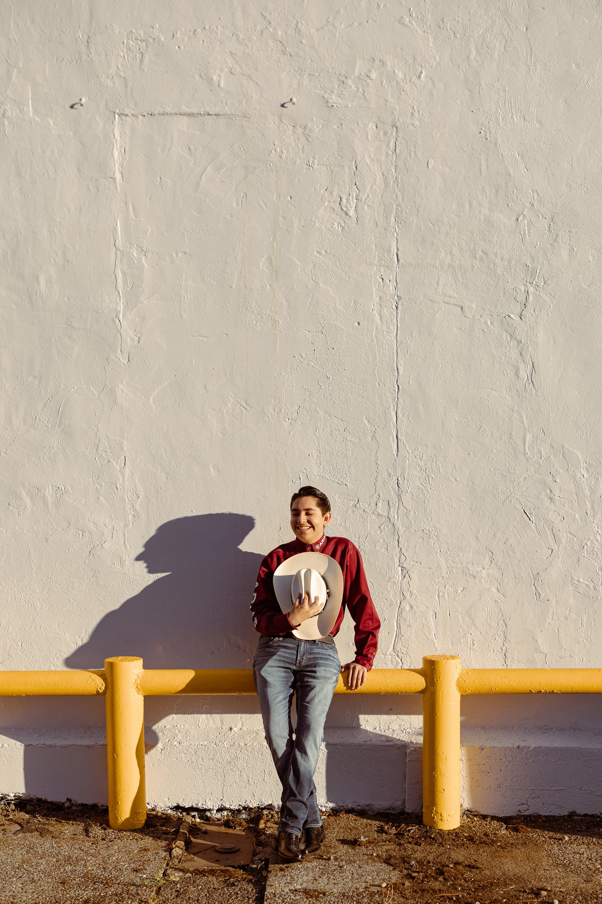 A young woman standing against a white textured wall and yellow safety barriers, holding a beige wide-brimmed hat, smiling, wearing a maroon jacket, gray jeans, and black boots. It is daytime, and her shadow is cast on the wall.