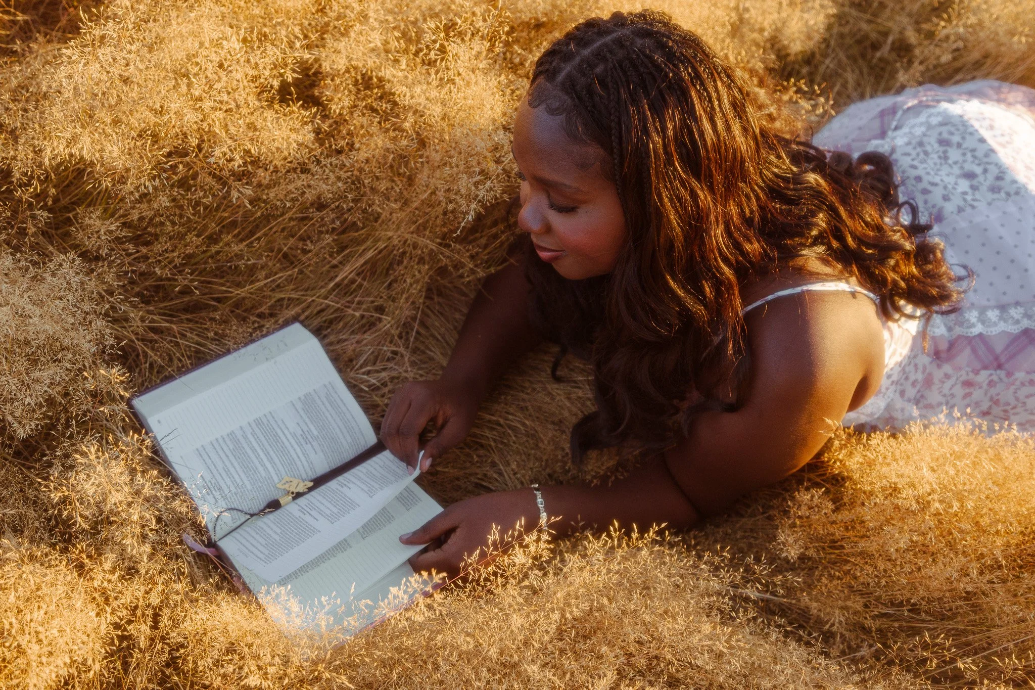 A woman with dark curly hair lying on her stomach in a field of tall, golden grass, reading a book with a ribbon bookmark during sunset.