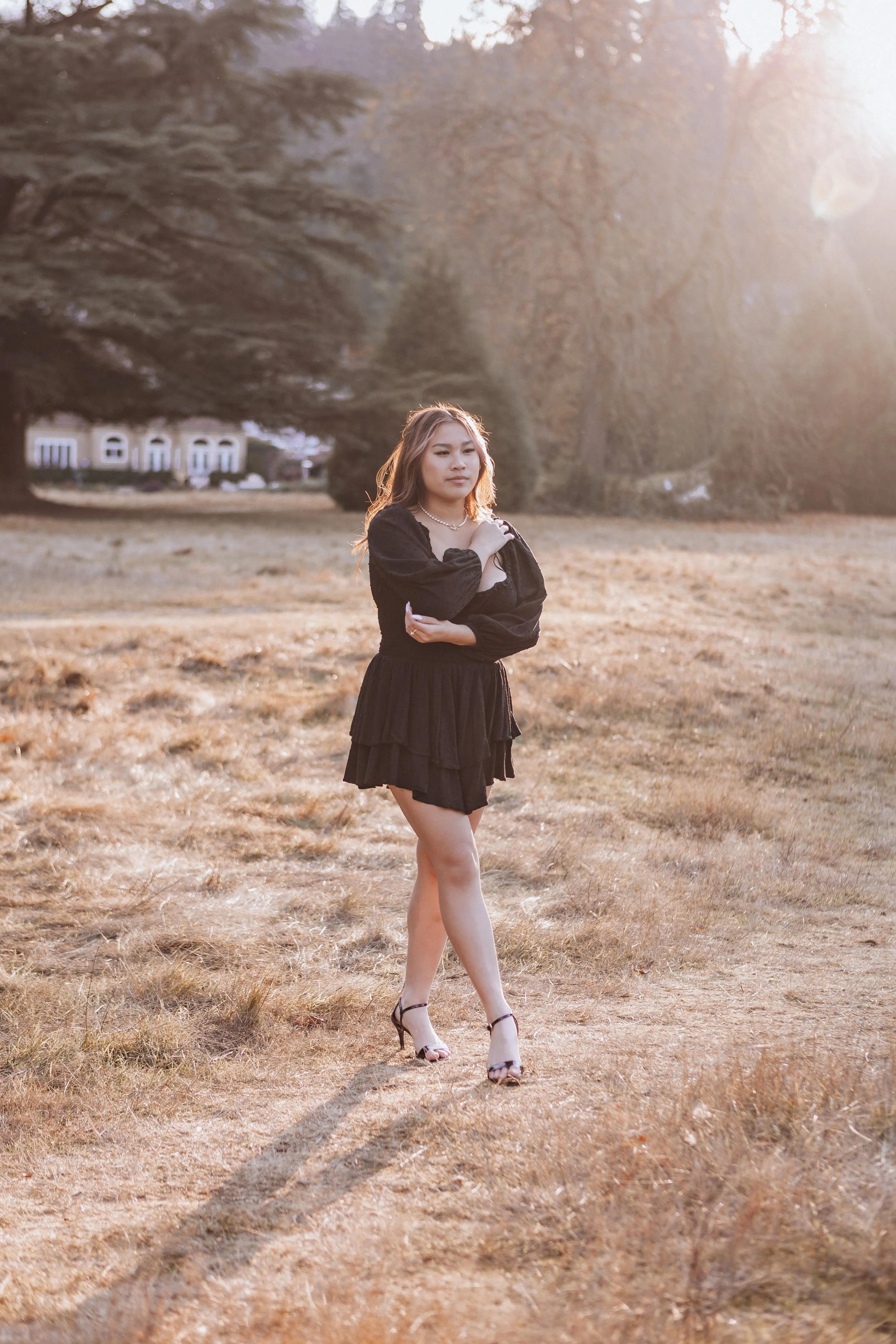 A woman in a black dress with puffed sleeves standing outdoors on dry grass with trees and houses in the background, illuminated by sunlight.