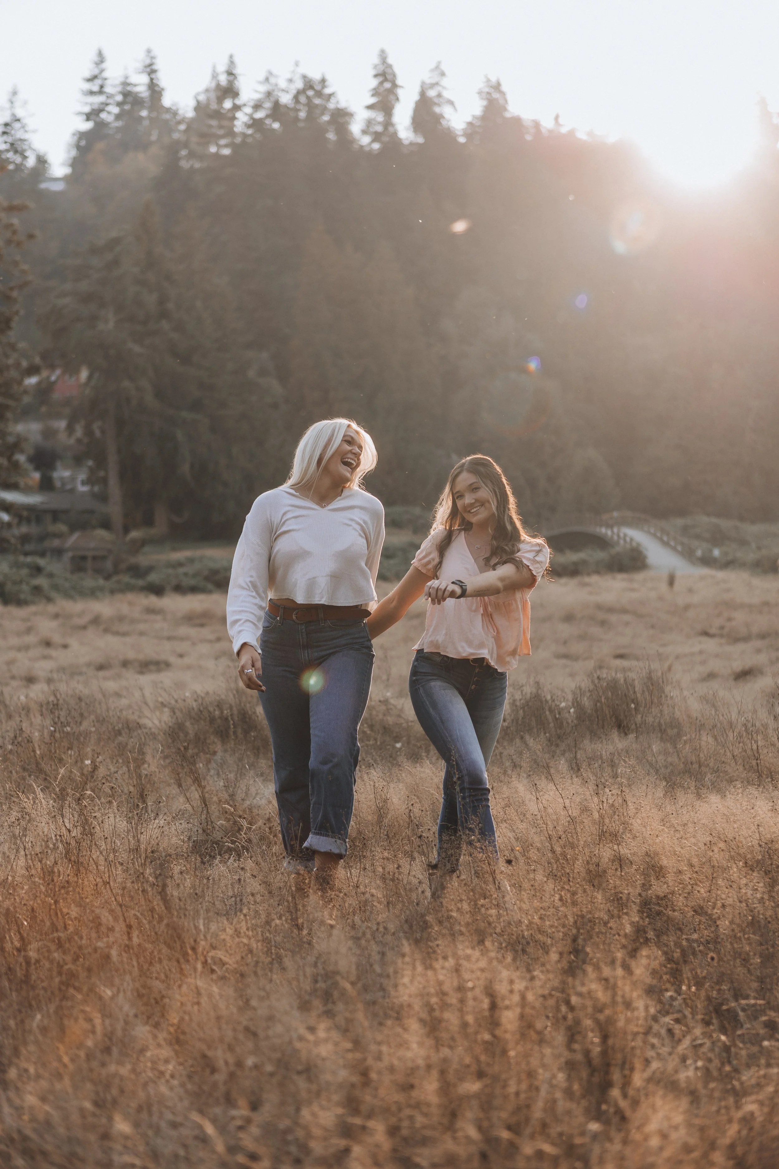 Two women walking through a field, smiling and looking at each other, with trees and a bridge in the background, sunlight shining brightly from the upper right corner.