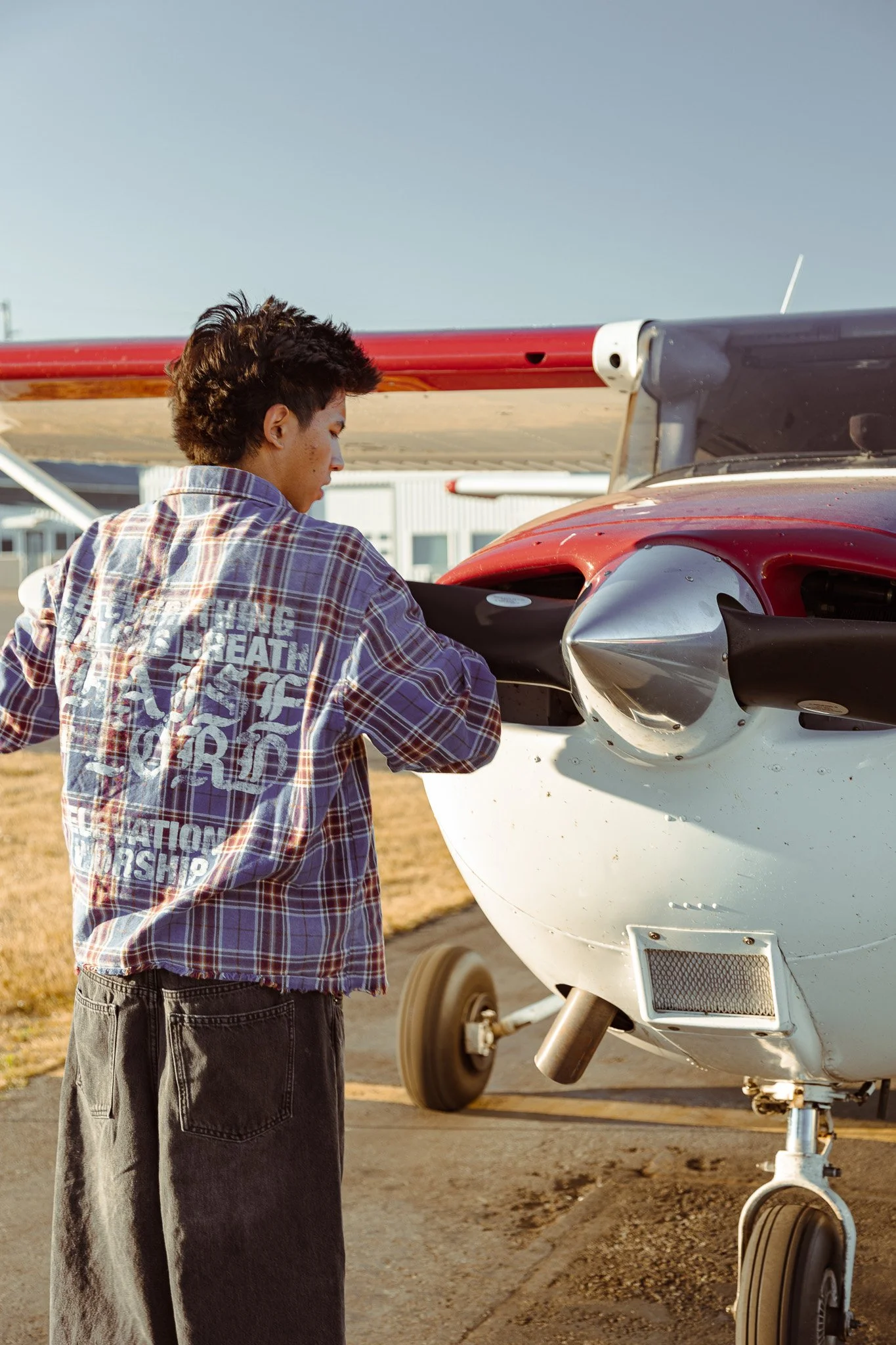 A young man with brown hair wearing a plaid shirt and dark jeans is standing next to a small private airplane, inspecting it outdoors under a clear sky.